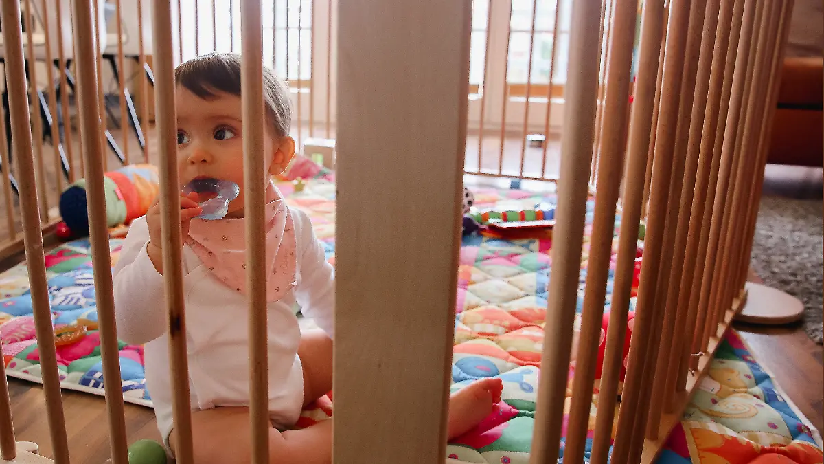 Ein Baby schaut durch die Gitter eines Laufstalls und kaut auf einem Beißring.