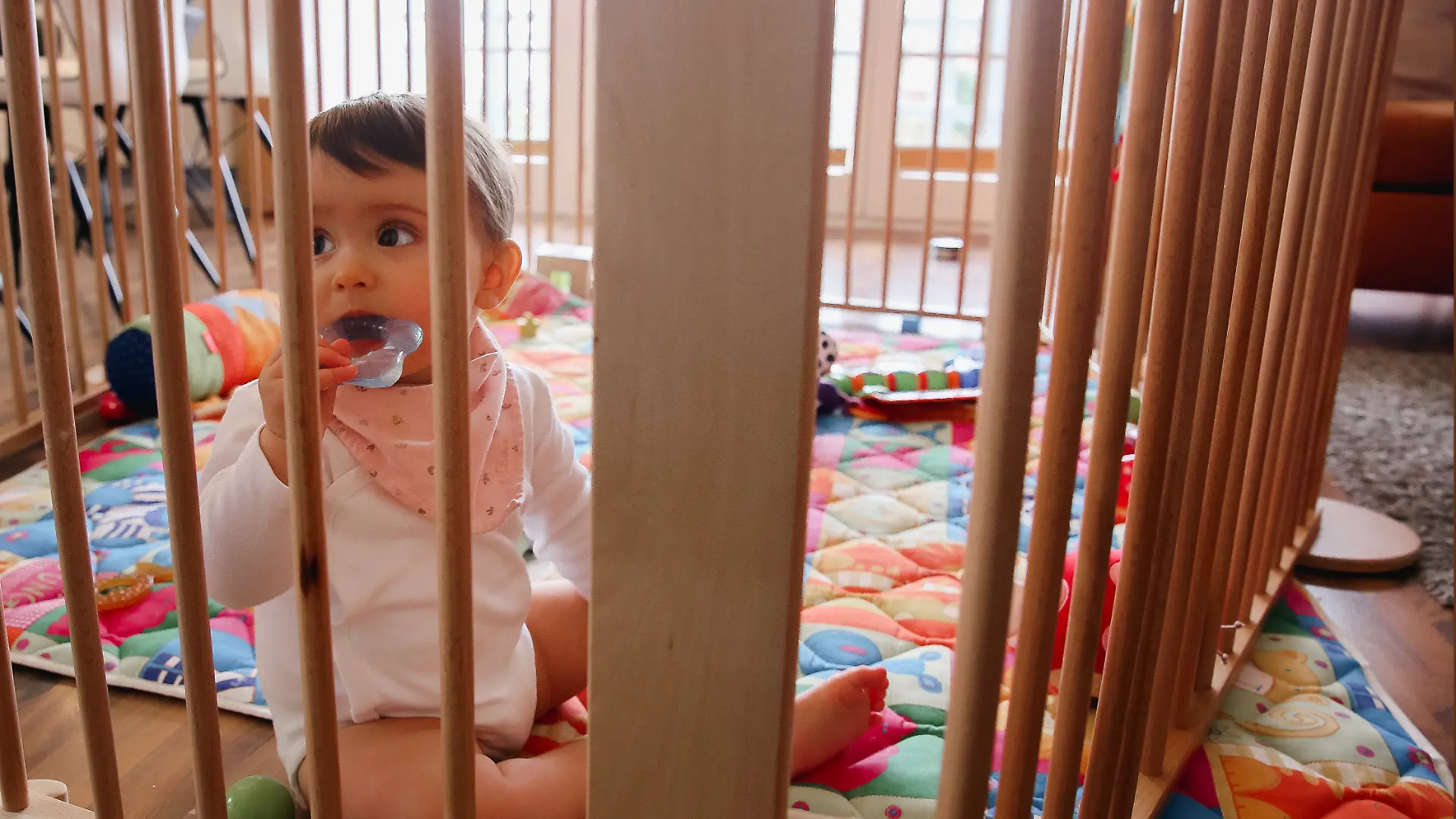 Ein Baby schaut durch die Gitter eines Laufstalls und kaut auf einem Beißring.
