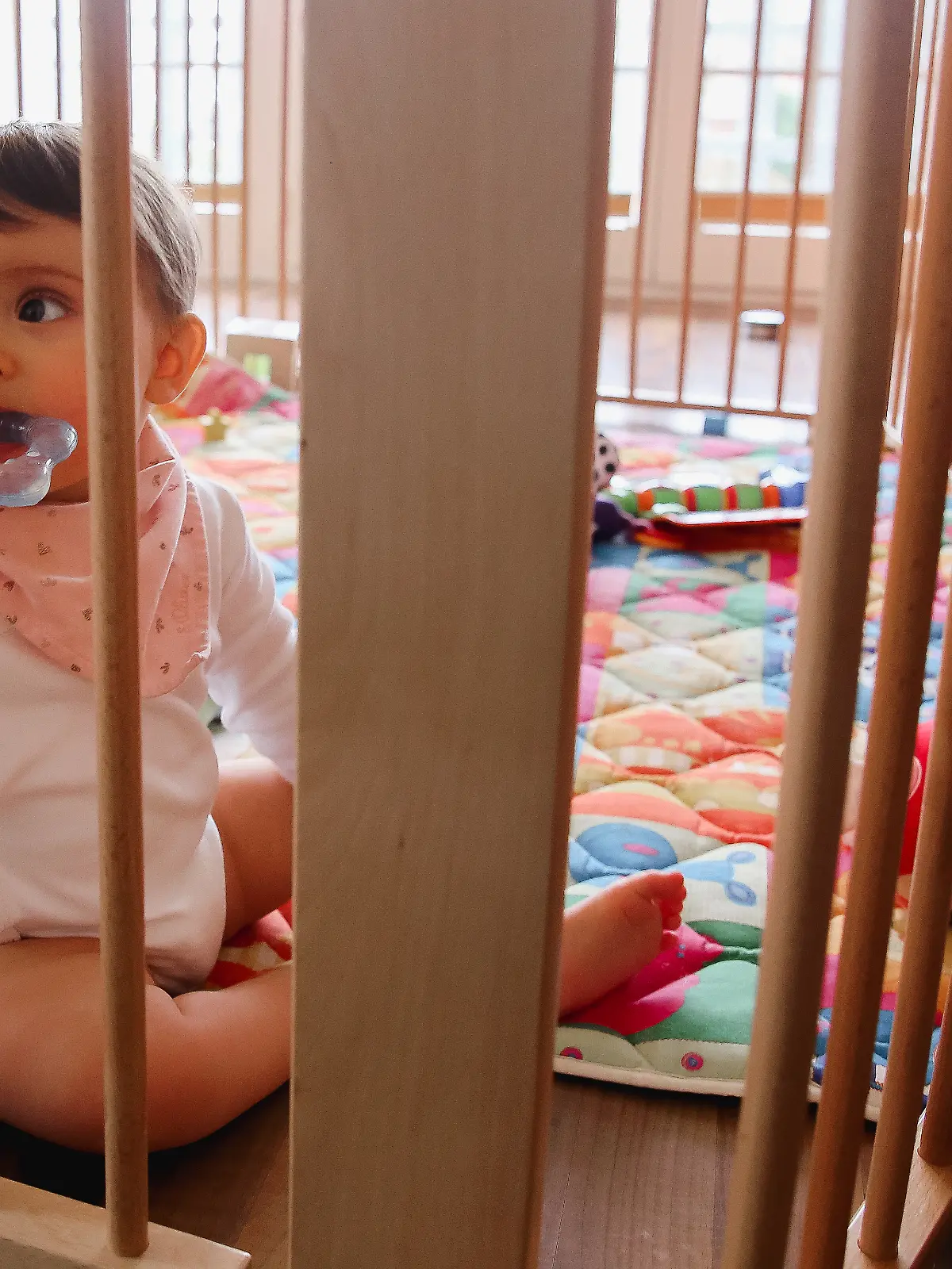 Ein Baby schaut durch die Gitter eines Laufstalls und kaut auf einem Beißring.