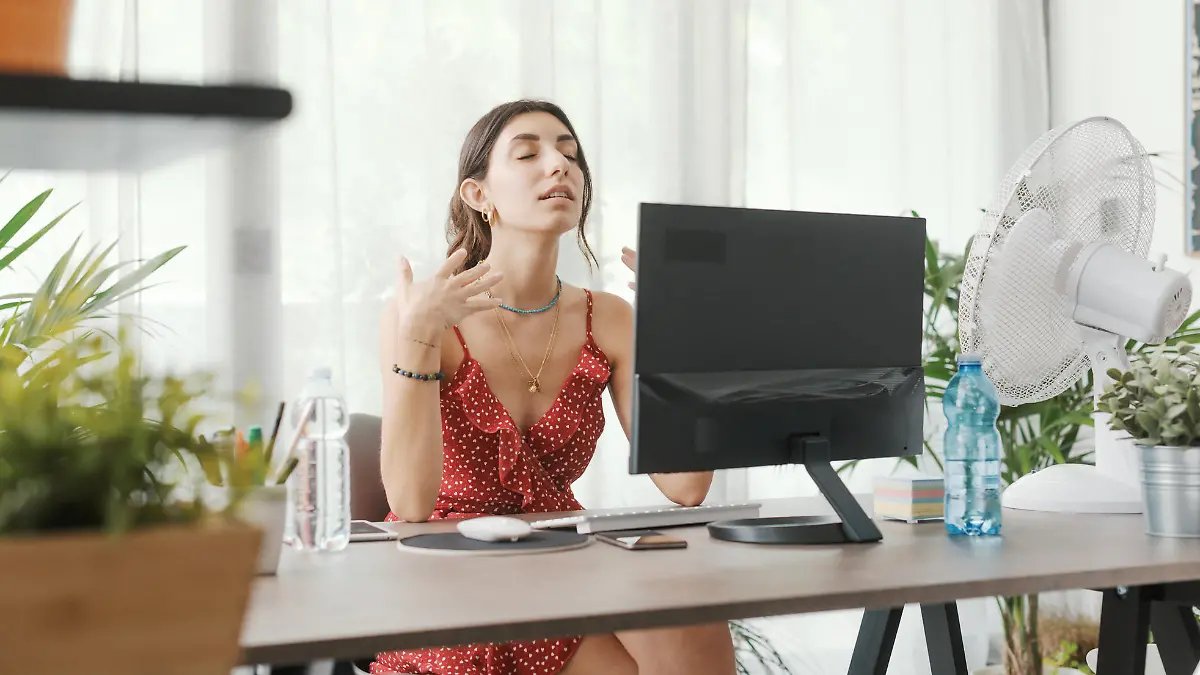 Woman sitting at desk at home during a summer heat wave, she is cooling herself with an electric fan