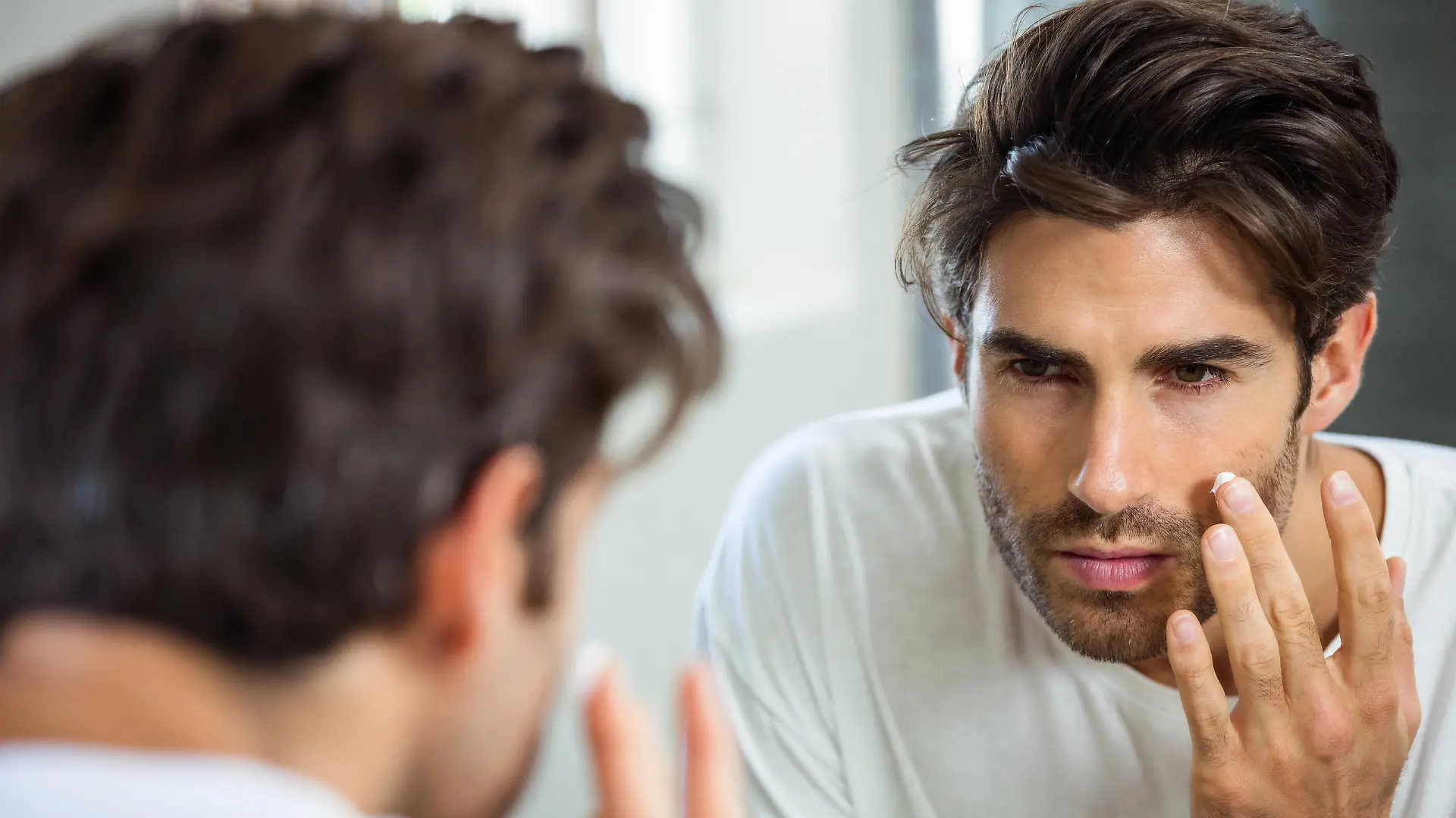 Reflection of young man in bathroom mirror applying moisturizer on his face