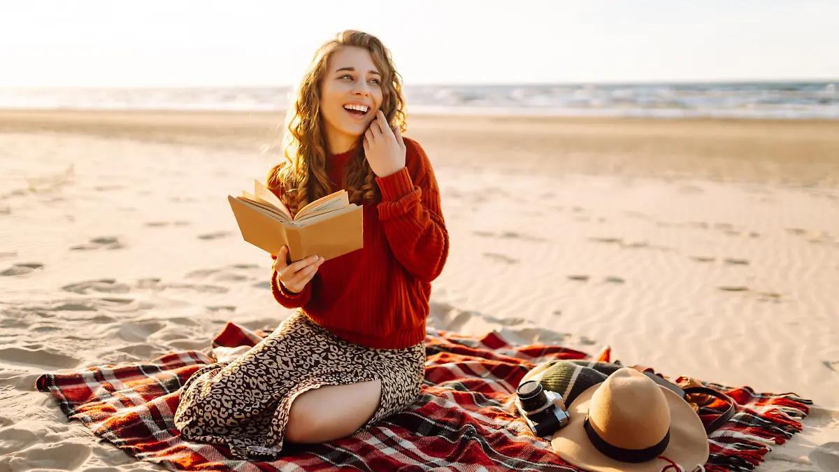 Beautiful woman sitting on the blanket reading a book at the beach picnic. The concept of relaxation, enjoyment, solitude with nature.