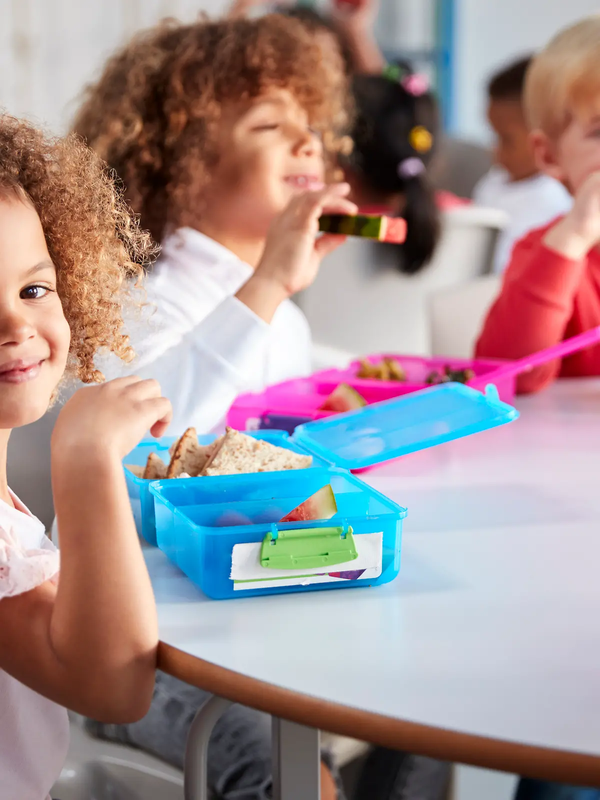 Kleines Mädchen mit lockigen Haaren sitzt im Kindergarten am Tisch vor ihrer Brotdose und lächelt in die Kamera.
