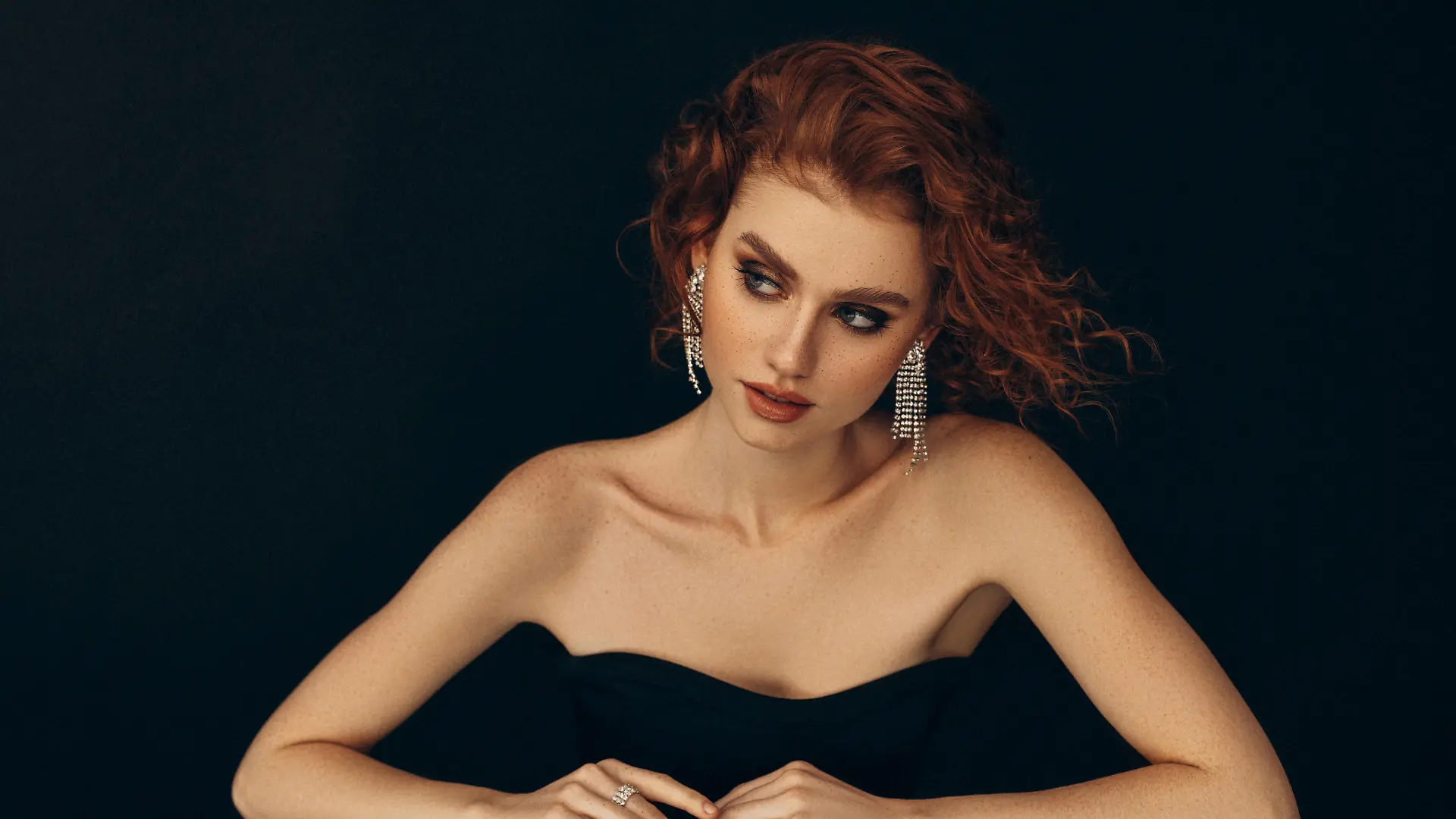 A striking portrait of a red-haired woman dressed in a chic black strapless dress. She sits gracefully against a dark background, with her arms elegantly positioned on a reflective surface. The woman's captivating gaze is directed slightly to the side, adding a sense of mystery to her poised demeanor. Her makeup is flawless, highlighting her natural beauty, and her look is completed with a pair of dazzling, statement earrings that catch the light. The contrast between her fair skin and the dark backdrop enhances the sophisticated and timeless quality of the image.