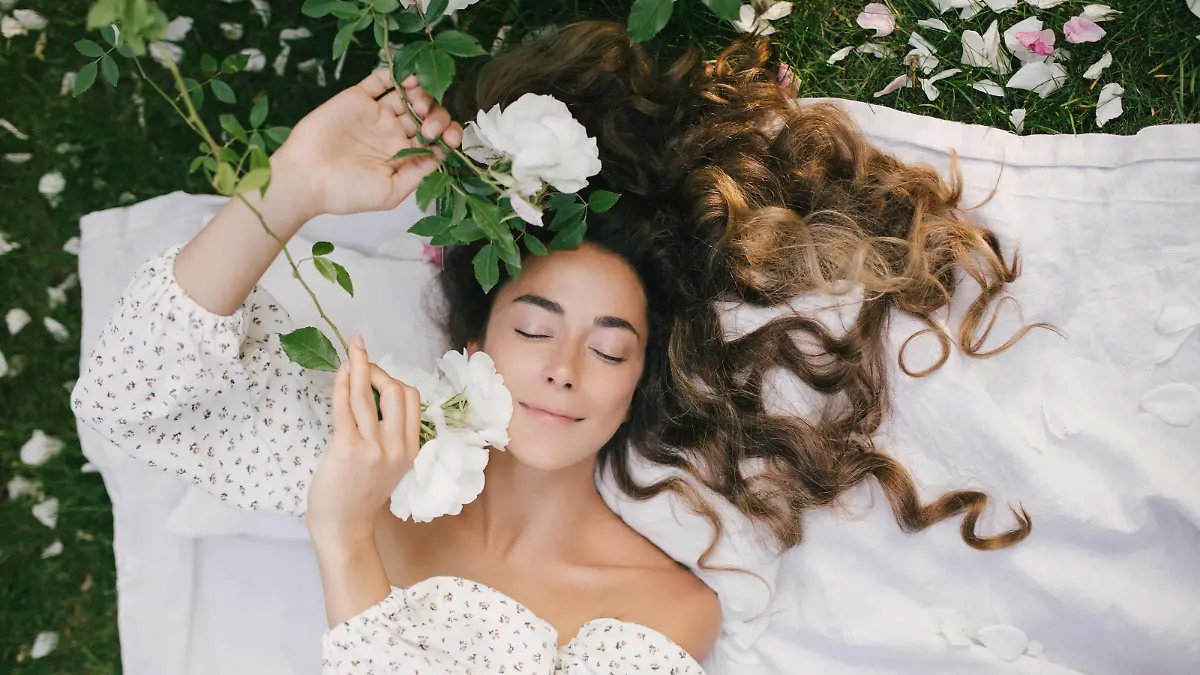 Young beautiful woman with long curly hair and perfect skin wearing white dress relaxing in blooming rose garden.
