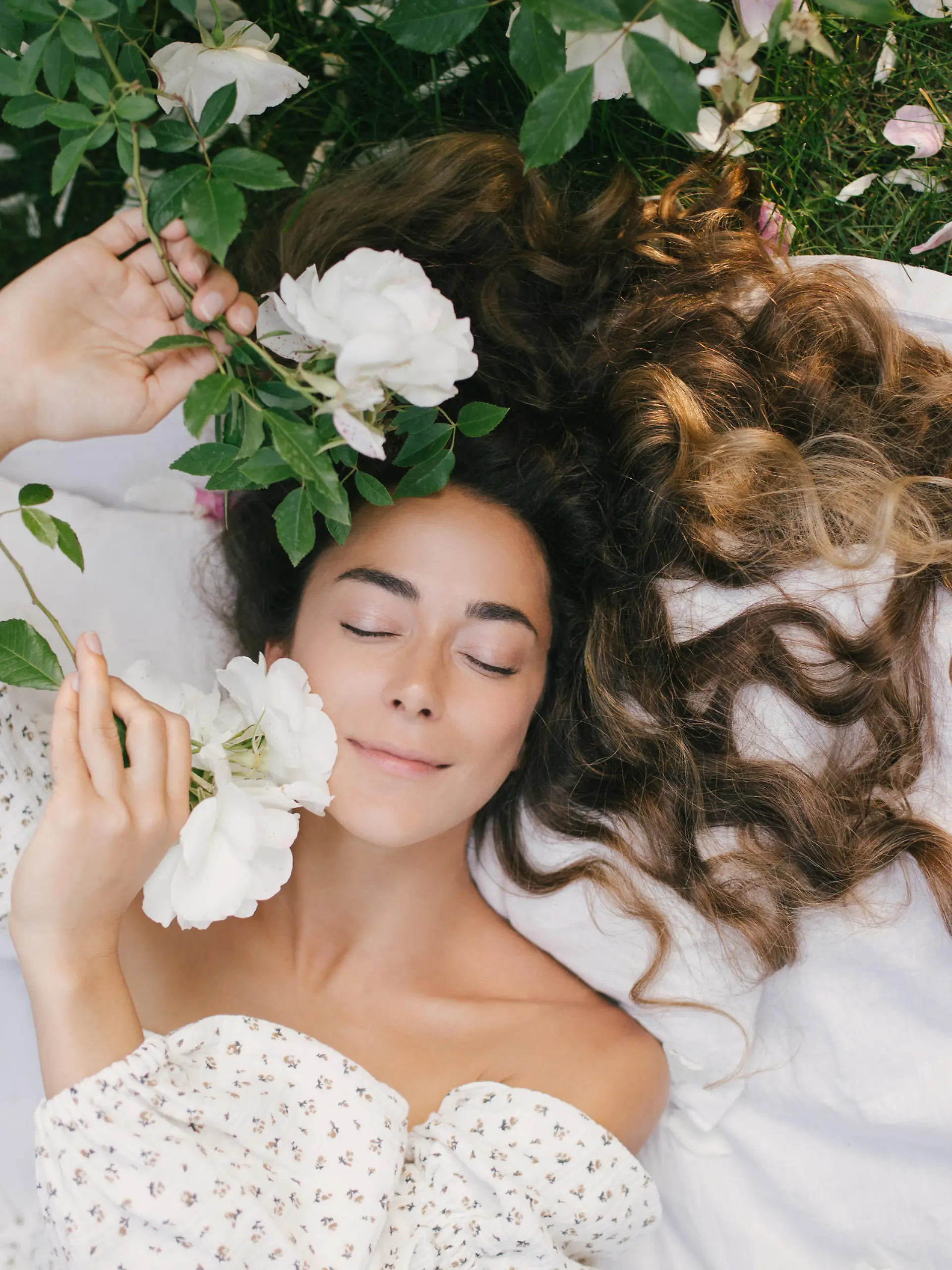 Young beautiful woman with long curly hair and perfect skin wearing white dress relaxing in blooming rose garden.