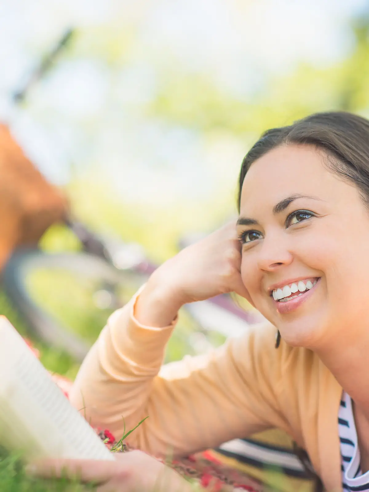 Lächelnde Frau liegt mit einem Buch auf einer Wiese und schaut in die Ferne.