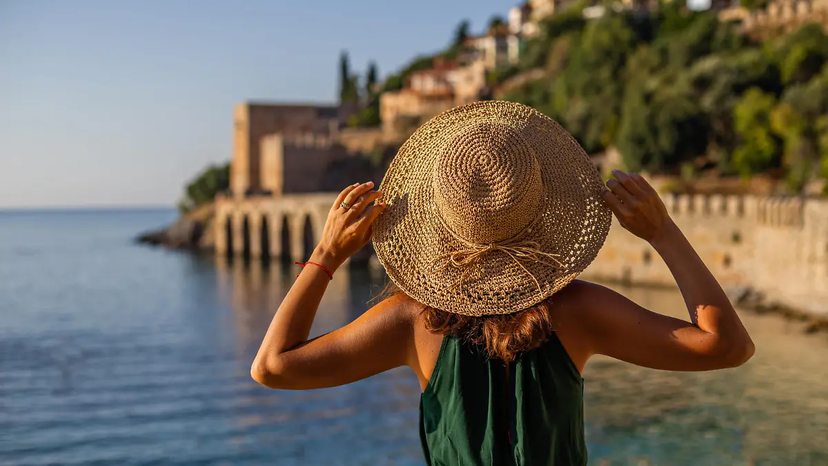 Horizontal backview portrait of a fit female wearing green top and straw hat and standing in front of Alanya castle wall and medieval shipyard