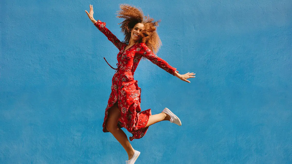 Young female model in red sundress jumping with joy. Beautiful young woman jumping over blue background.