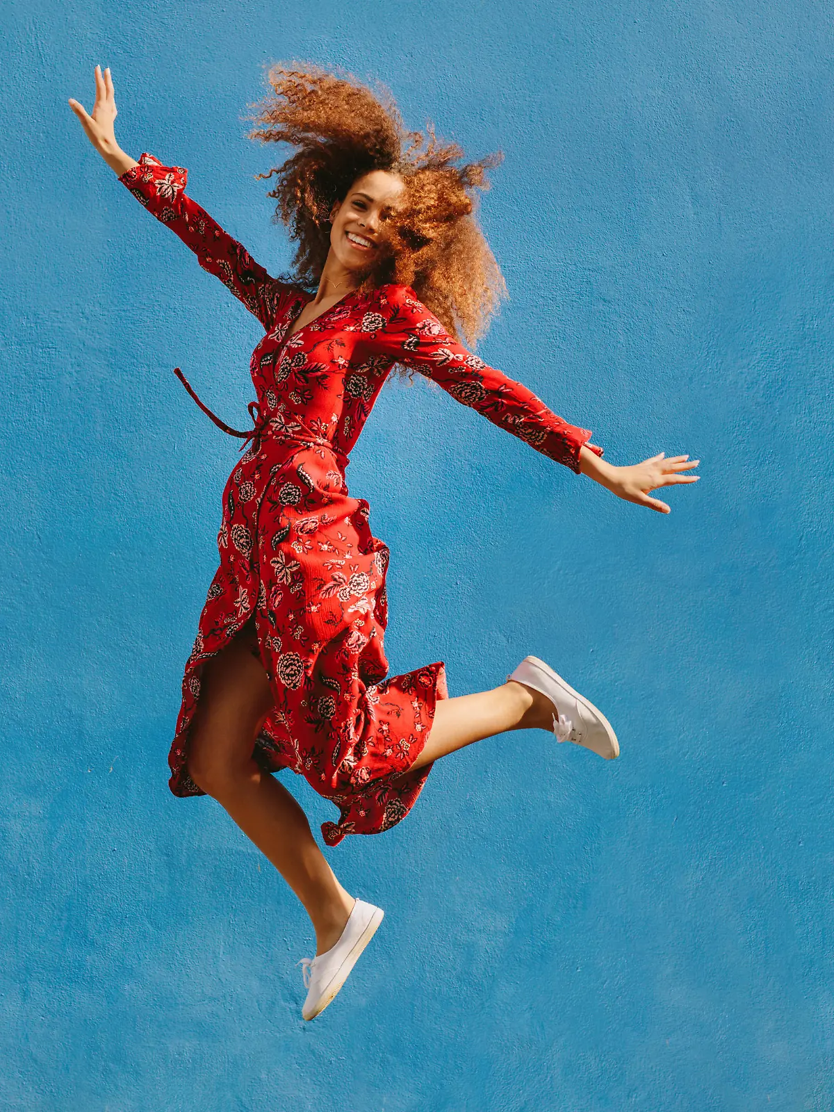 Young female model in red sundress jumping with joy. Beautiful young woman jumping over blue background.
