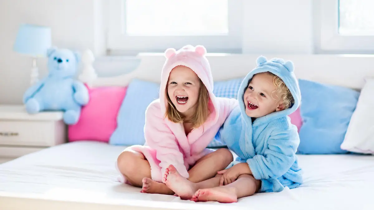 Happy laughing kids, boy and girl in soft bathrobe after bath play on white bed with blue and pink pillows in sunny bedroom. Child in clean and dry towel. Wash, infant hygiene, health and skin care.