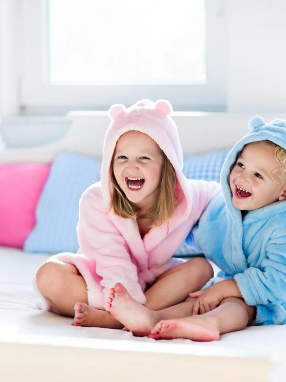 Happy laughing kids, boy and girl in soft bathrobe after bath play on white bed with blue and pink pillows in sunny bedroom. Child in clean and dry towel. Wash, infant hygiene, health and skin care.