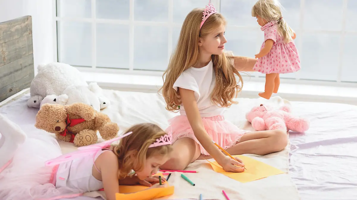 Future artists. Good-looking girls drawing different pictures. Elder sister is painting her doll and smiling. Toys are on the big bed