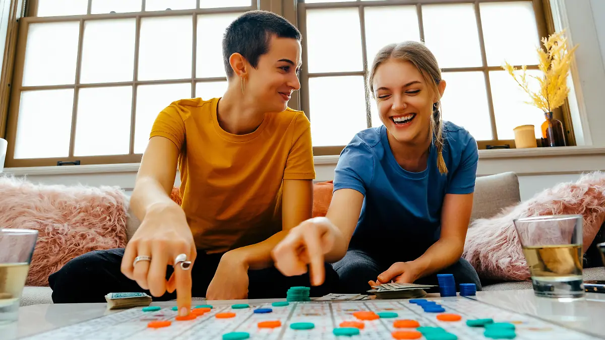 Two young women sitting on couch playing board game with counters, laughing. Happy couple bonding over an indoor leisure activity, smiling and sitting together at a table.
