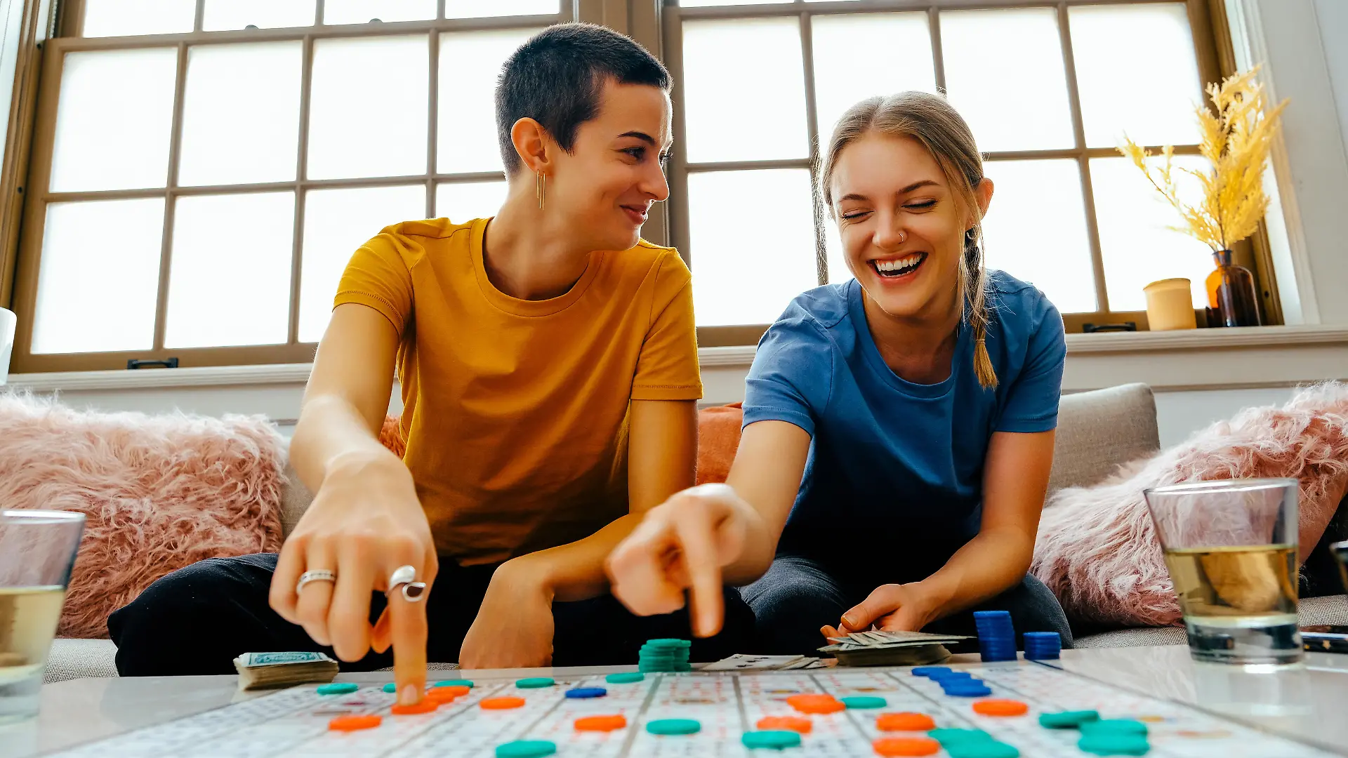 Two young women sitting on couch playing board game with counters, laughing. Happy couple bonding over an indoor leisure activity, smiling and sitting together at a table.