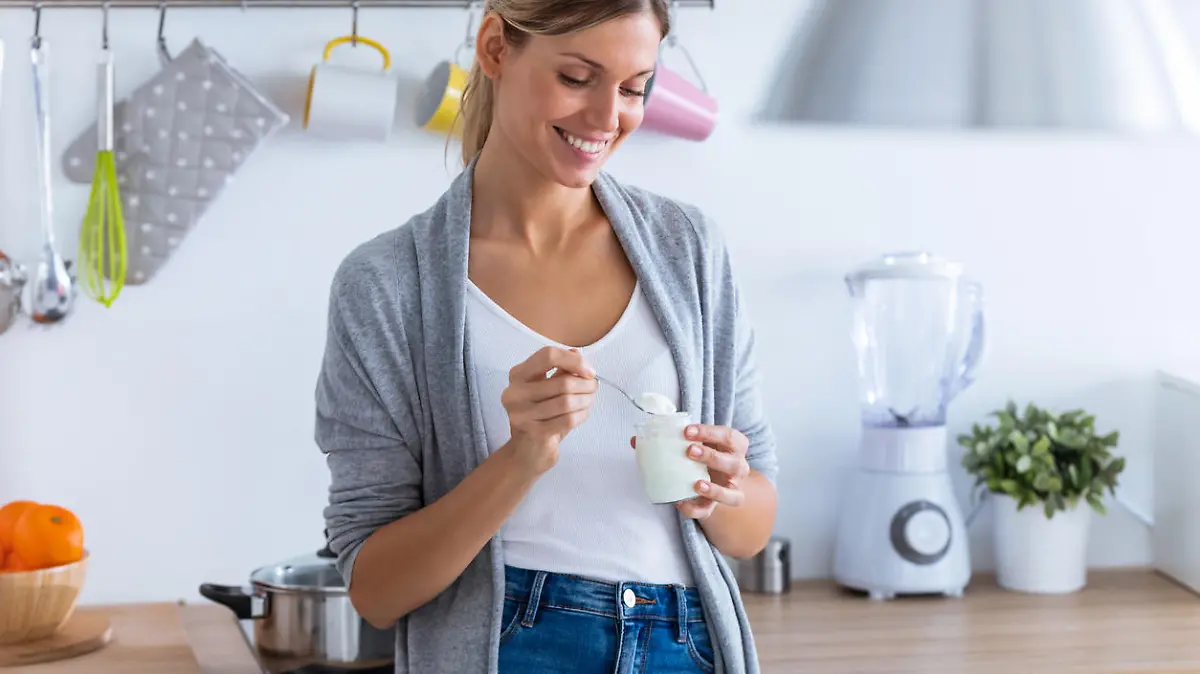 Shot of pretty young woman eating yogurt while standing in the kitchen at home.