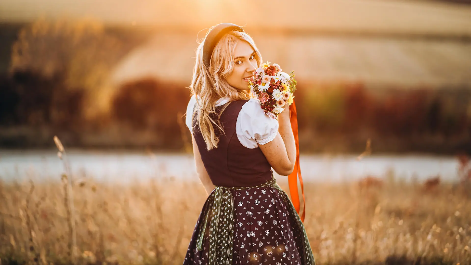 Pretty blonde girl in dirndl, traditional festival dress, standing outdoors in the field at sunset time, holding bouquet of a field flowers. Beer Fest, St. Patrick’s day, international beer day concept