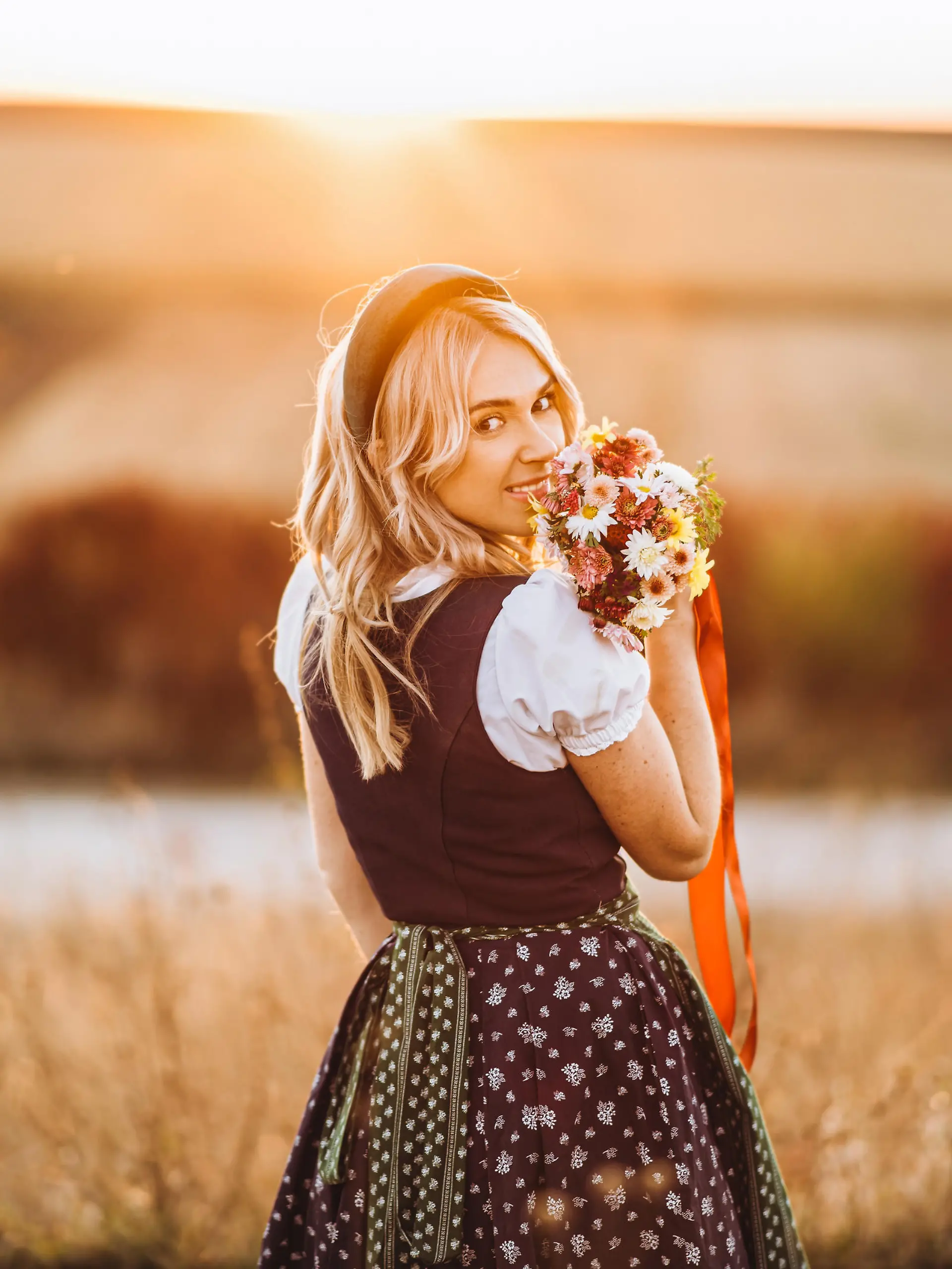 Pretty blonde girl in dirndl, traditional festival dress, standing outdoors in the field at sunset time, holding bouquet of a field flowers. Beer Fest, St. PatrickÃ¢Â€Â™s day, international beer day concept