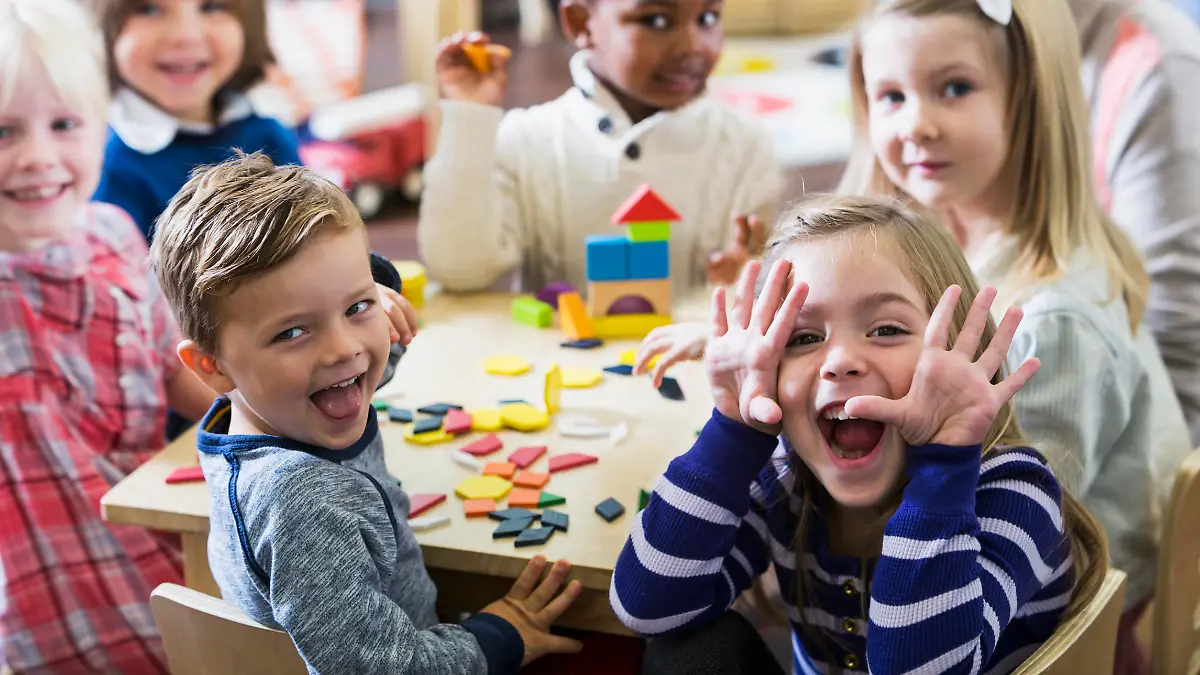 A multiracial group of preschoolers or kindergarteners having fun in the classroom. Six children are sitting around a little wooden table playing with colorful wooden block and geometric shapes. The playful little girl in the foreground is making a silly face at the camera.