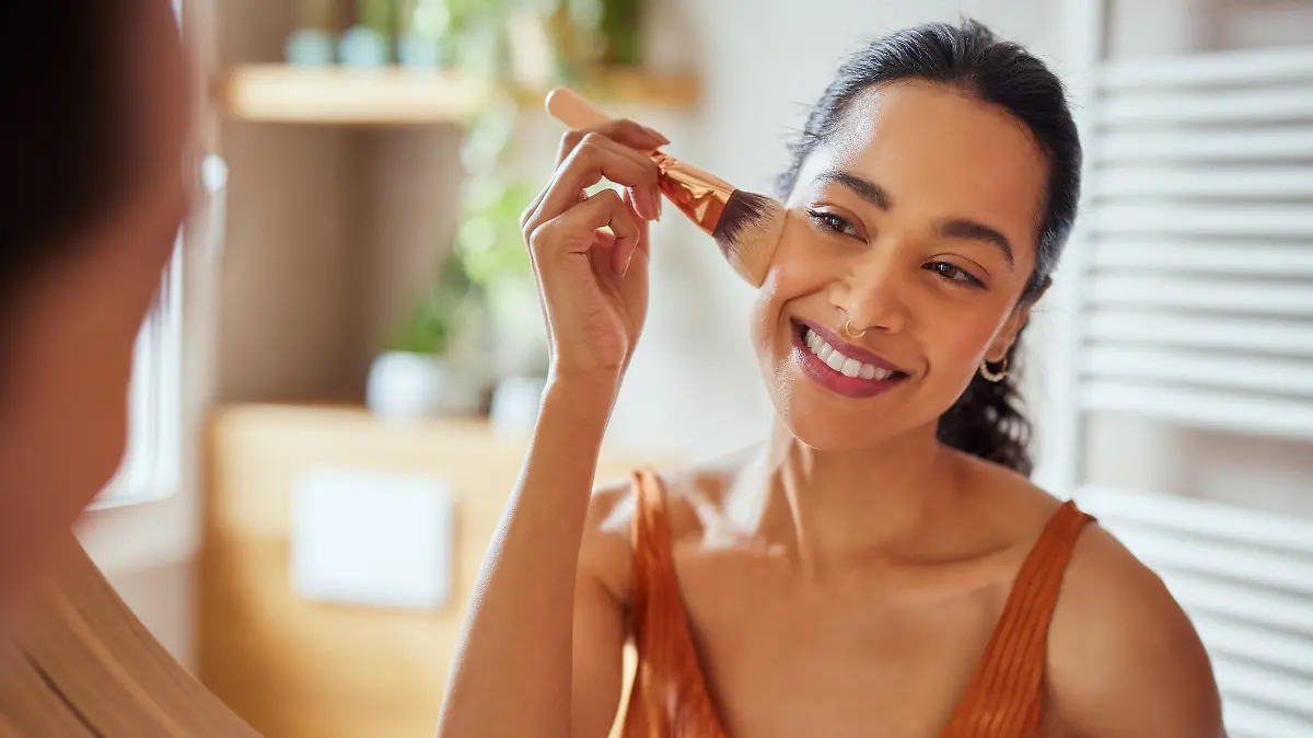 Beautiful girl applying makeup using powder brush before going to work. Healthy latin woman looking in the mirror and applying cosmetic with a big brush. Young woman looking in the mirror and applying foundation or blusher on her face.