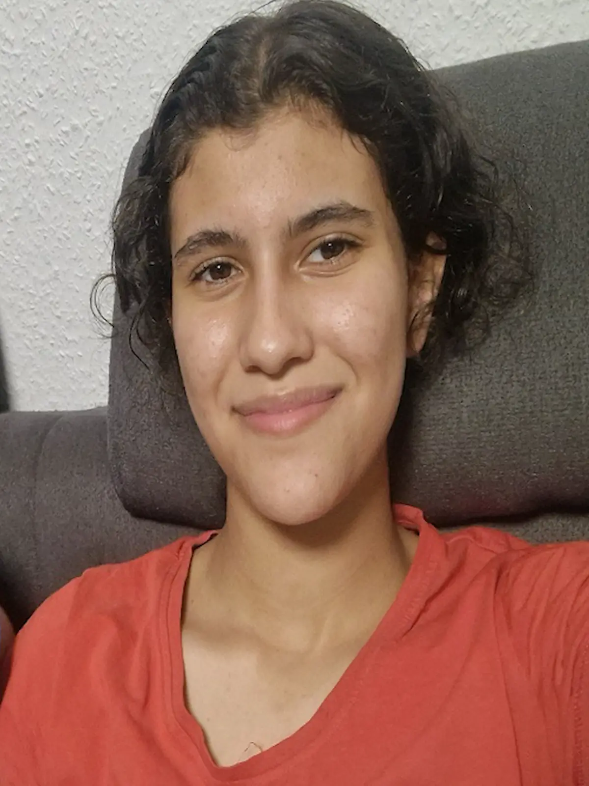 Ein Mädchen mit braunen Haaren und rotem T-Shirt macht ein Selfie auf einer Couch.