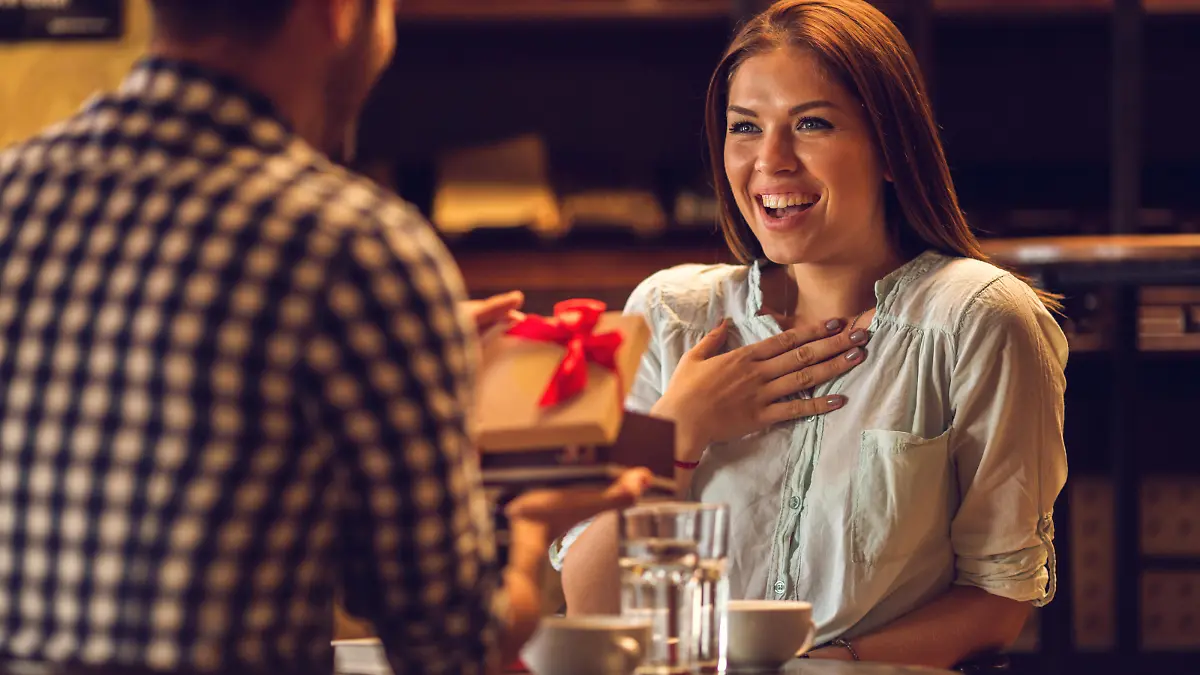 Young happy woman in disbelief receiving a present from her boyfriend while sitting in a cafe.