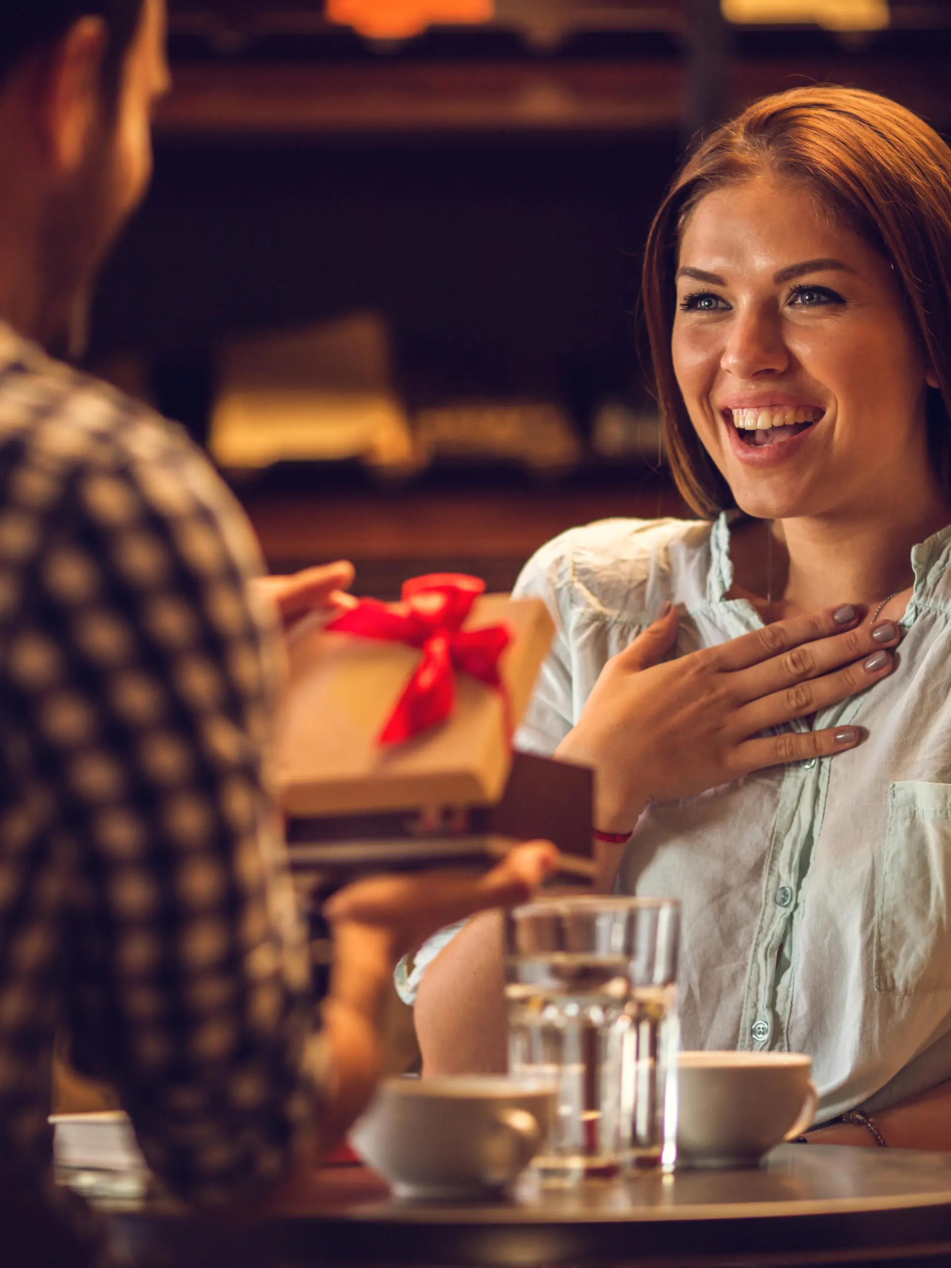 Young happy woman in disbelief receiving a present from her boyfriend while sitting in a cafe.
