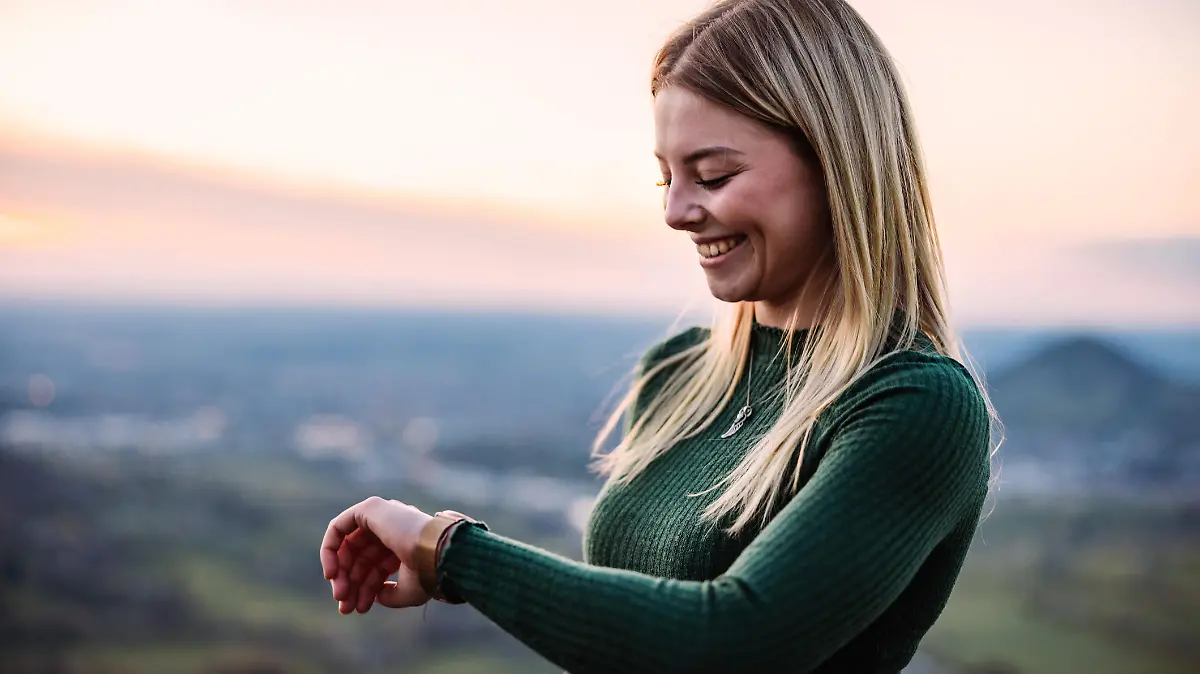 Young blonde millennial generation Girl standing outdoors in the nature during sunset looking down to her smart watch, checking messages, news and fitness data of his walk - hike. South German Mountain Range in the background. Backlit from Dusk - Sunset Sun. Real People Millennial Generation Outdoors Modern Technology Fitness Scene.