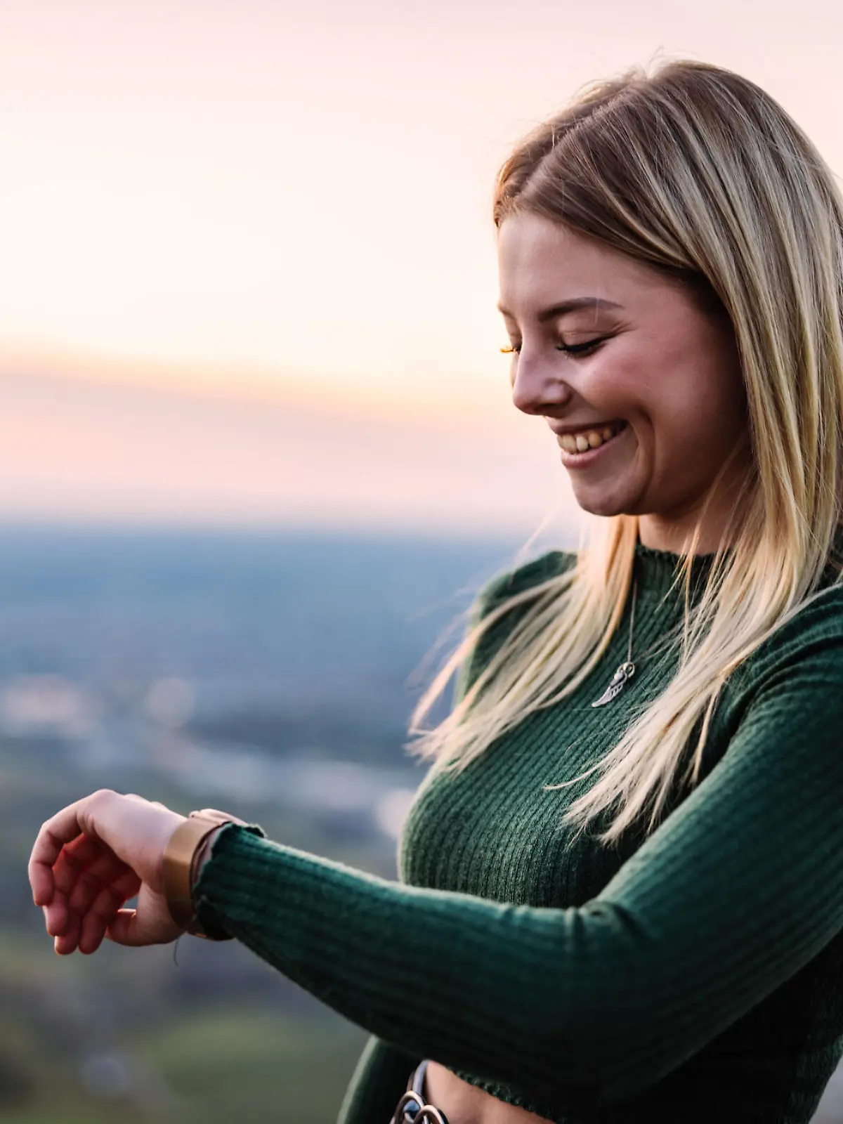 Young blonde millennial generation Girl standing outdoors in the nature during sunset looking down to her smart watch, checking messages, news and fitness data of his walk - hike. South German Mountain Range in the background. Backlit from Dusk - Sunset Sun. Real People Millennial Generation Outdoors Modern Technology Fitness Scene.