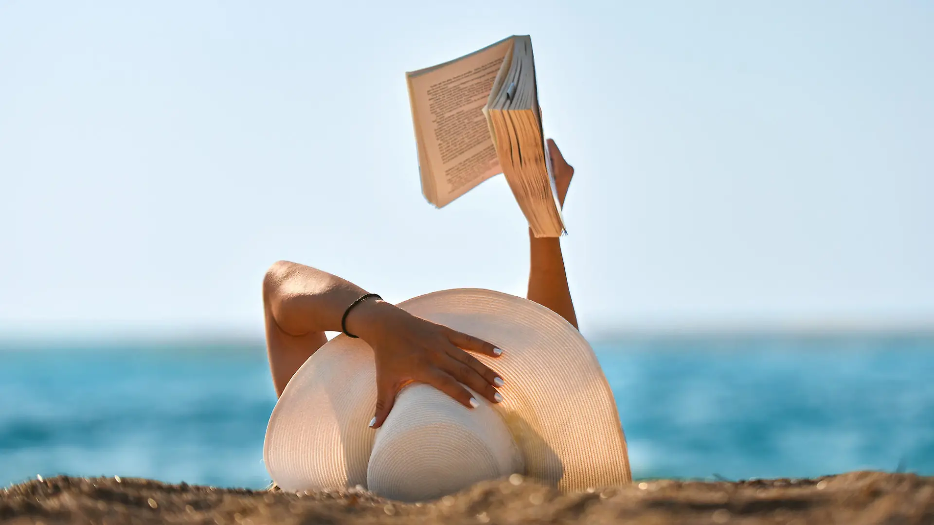 Frau mit Sonnenhut liest Buch am Strand