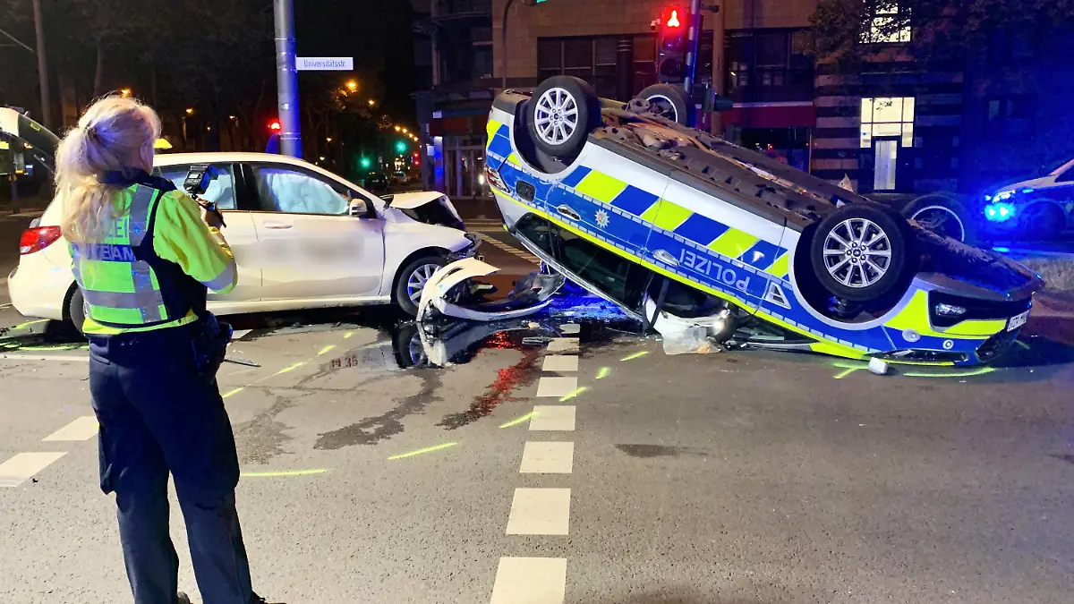07.05.2022, Nordrhein-Westfalen, Köln: Eine polizistin betrachtet die Unfallstelle. Auf dem Weg zu einem Einsatz ist ein Streifenwagen in Köln in der Nacht auf Samstag mit einem Taxi kollidiert - dabei sind sechs Menschen verletzt worden. Wie ein Sprecher der Polizei mitteilte, waren zwei Beamte ursprünglich zu einer Schlägerei gerufen worden und bogen mit ihrem Wagen links ab, als ihnen ein Taxi entgegenkam. Der Taxifahrer konnte wohl nicht mehr rechtzeitig bremsen und fuhr seitlich in den Streifenwagen. Dieser überschlug sich und blieb auf dem Dach liegen. Foto: Daniel Evers/WupperVideo/dpa +++ dpa-Bildfunk +++