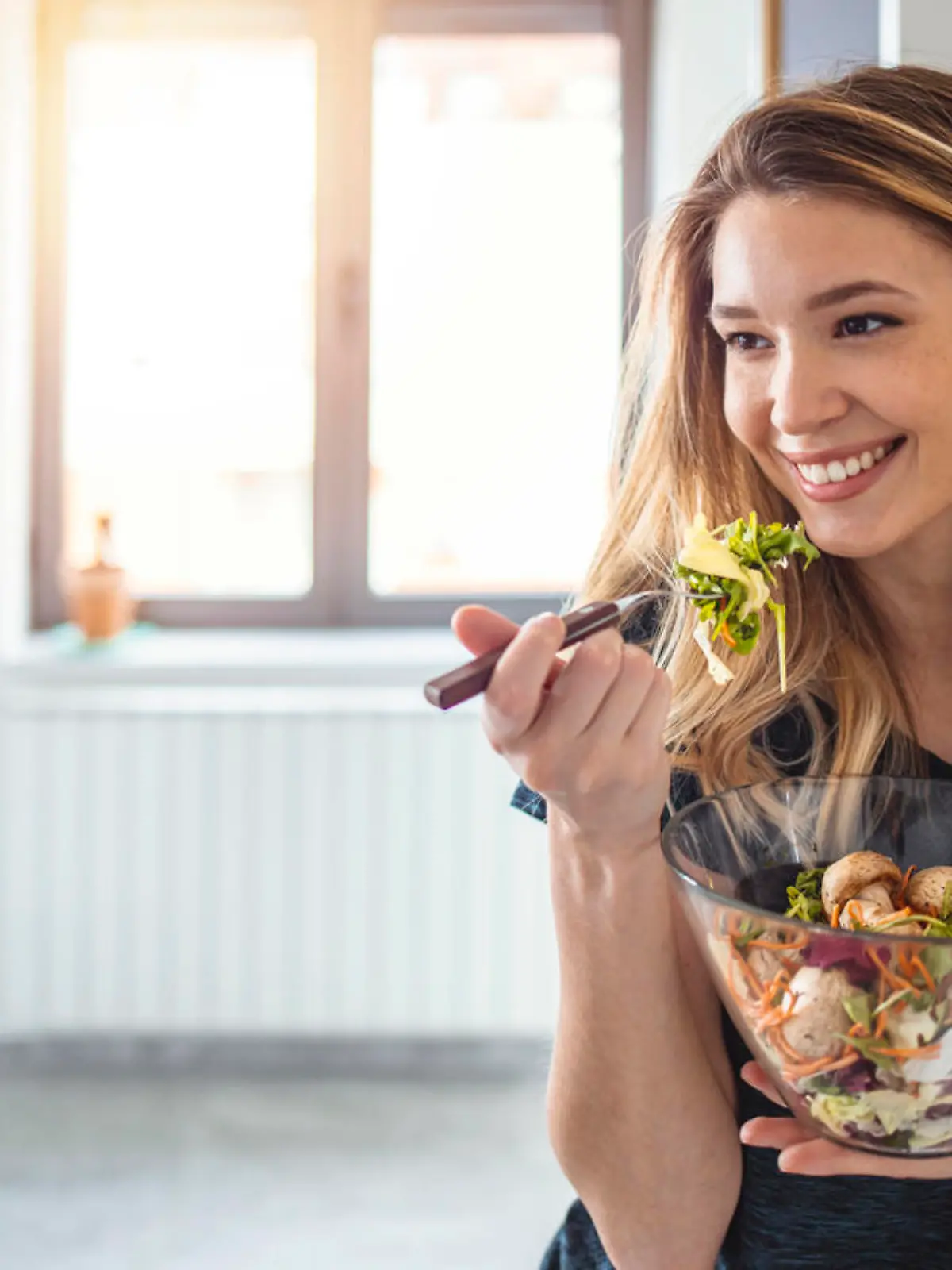 Starting day with healthy food. Beautiful young woman eating salad and looking away while standing in kitchen at home, looking away.