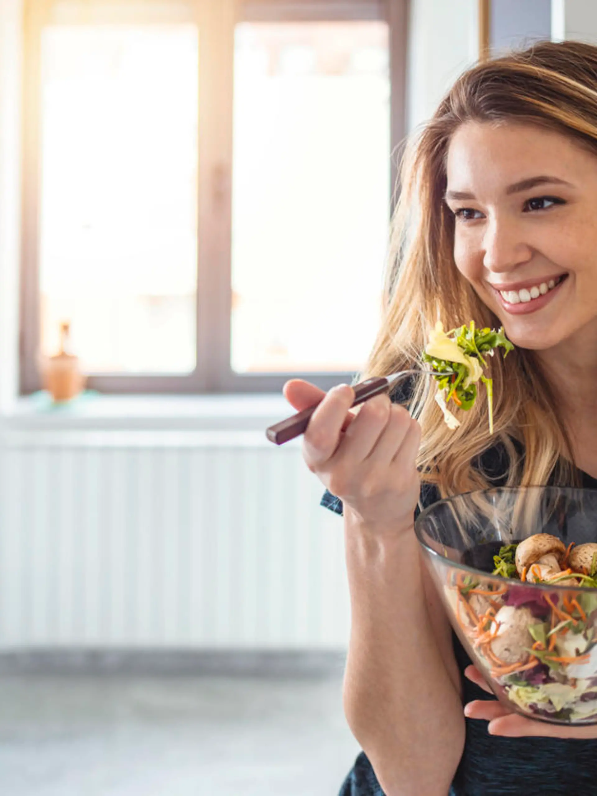Starting day with healthy food. Beautiful young woman eating salad and looking away while standing in kitchen at home, looking away.