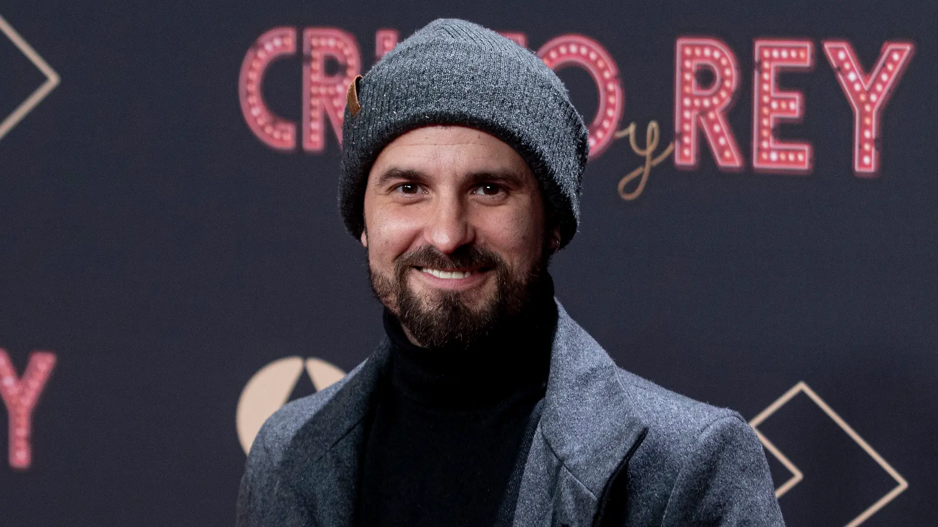 Schauspieler Julián Ortega trägt eine Wollmütze und einen grauen Anzug vor einer Werbewand. Der Schauspieler starb überraschend im August 2024. 