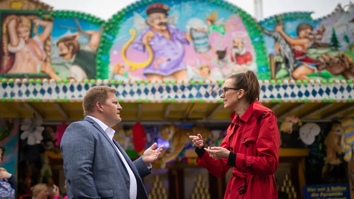 05.05.2022, Baden-Württemberg, Stuttgart: Mark Roschmann (l), Vorsitzender des Stuttgarter Schaustellerverbandes, spricht auf dem Frühlingsfest mit Stadträtin Jitka Sklenarova (Bündnis 90/Die Grünen) über das Thema Sexismus an einigen Schaustellerbuden - Wie bei dieser im Hintergrund mit freizügigen Frauen oben links und rechts. Foto: Christoph Schmidt/dpa - ACHTUNG: Nur zur redaktionellen Verwendung im Zusammenhang mit der aktuellen Berichterstattung und Verwendung nur im vollen Format. +++ dpa-Bildfunk +++