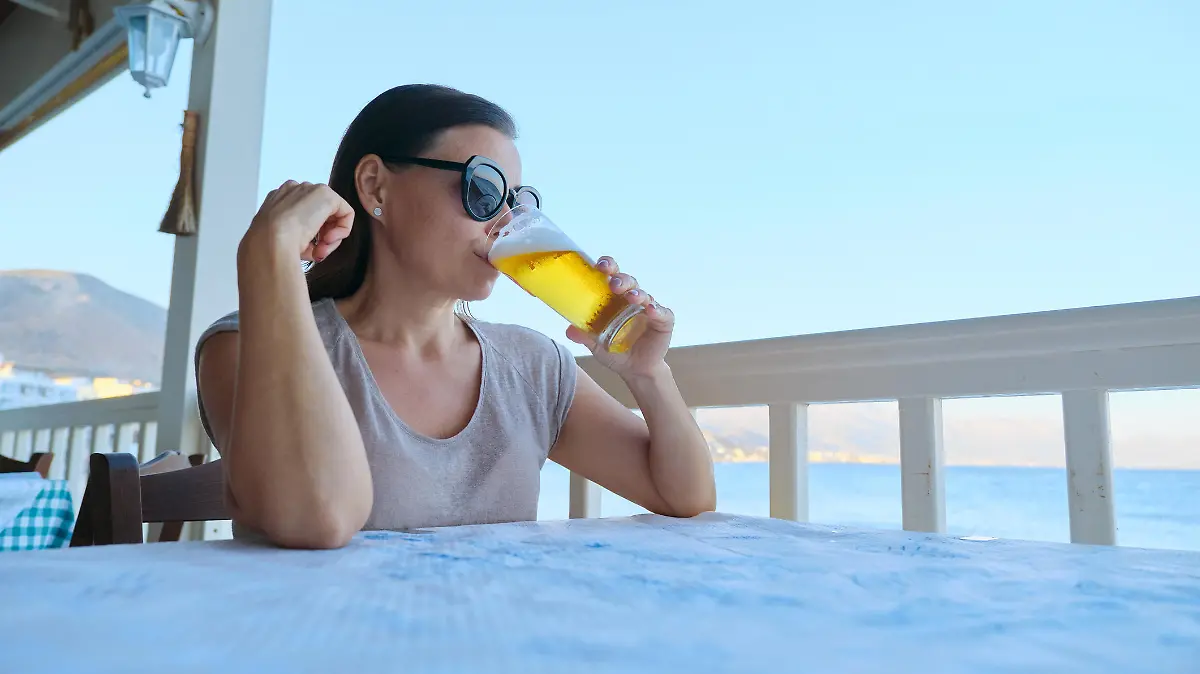 Mature beautiful woman resting sitting in sea cafe drinking cold glass of beer. Summer vacation, female enjoys sea sunset landscapes and delicious drink, copy space