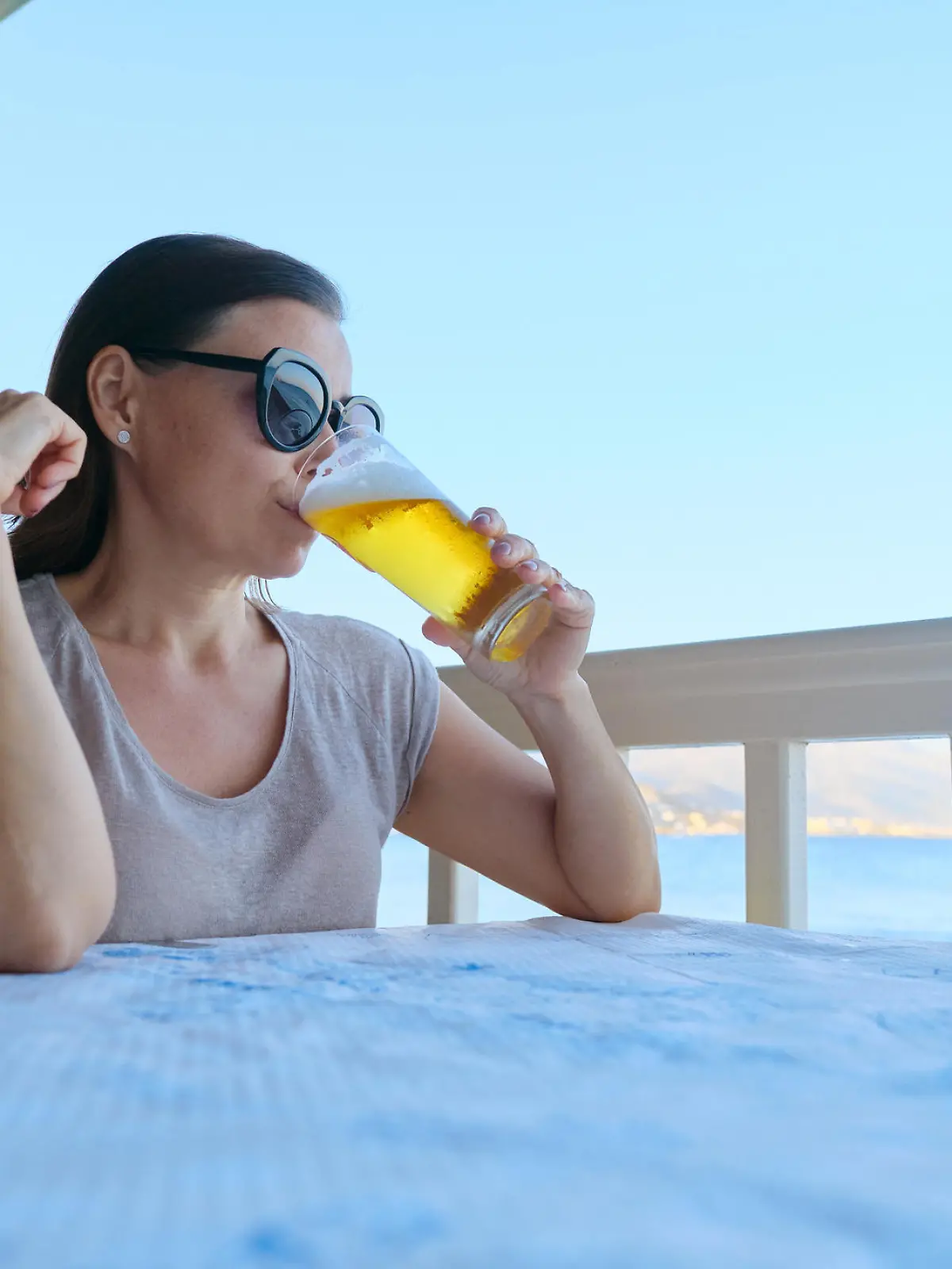 Mature beautiful woman resting sitting in sea cafe drinking cold glass of beer. Summer vacation, female enjoys sea sunset landscapes and delicious drink, copy space