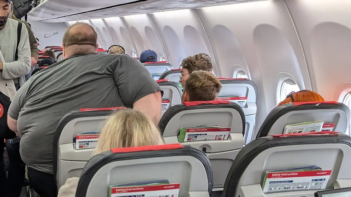 In einem Flugzeug versucht ein extrem übergewichtiger Mann sich auf einen Sitz zu setzen. Der Sitz ist deutlich zu klein, er engt damit den Gang und den Passagier neben sich ein.