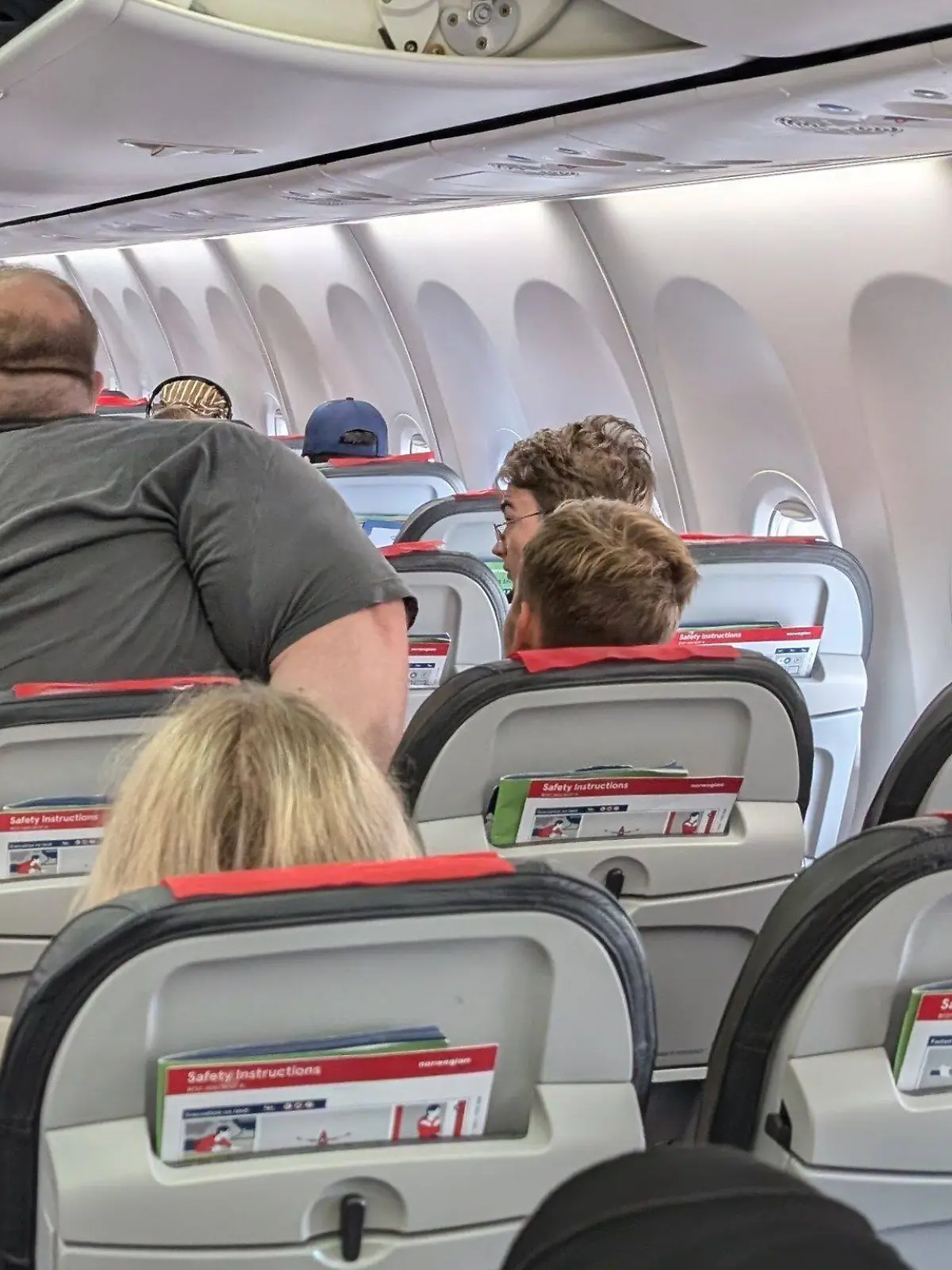 In einem Flugzeug versucht ein extrem übergewichtiger Mann sich auf einen Sitz zu setzen. Der Sitz ist deutlich zu klein, er engt damit den Gang und den Passagier neben sich ein.