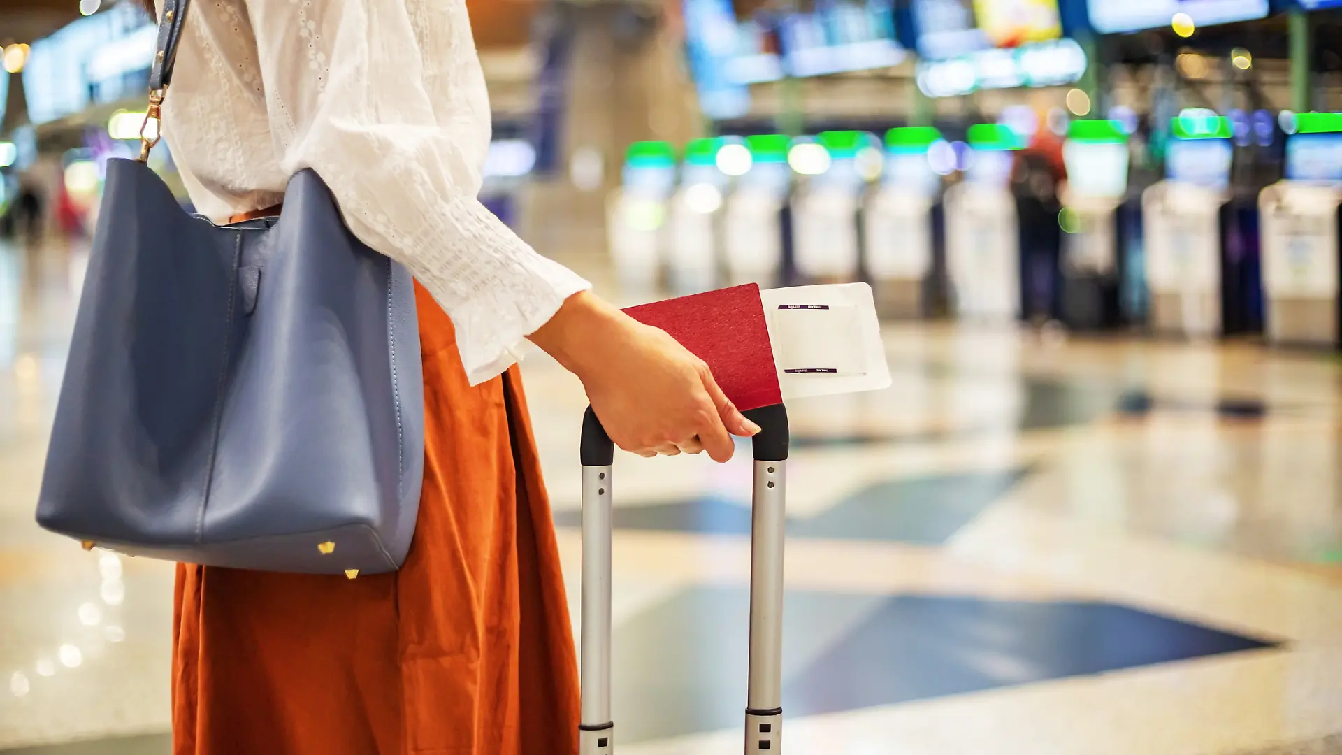 Eine Person steht im Foyer eines Flughafens mit einem kleinen Koffer und einer Handtasche über der Schulter.