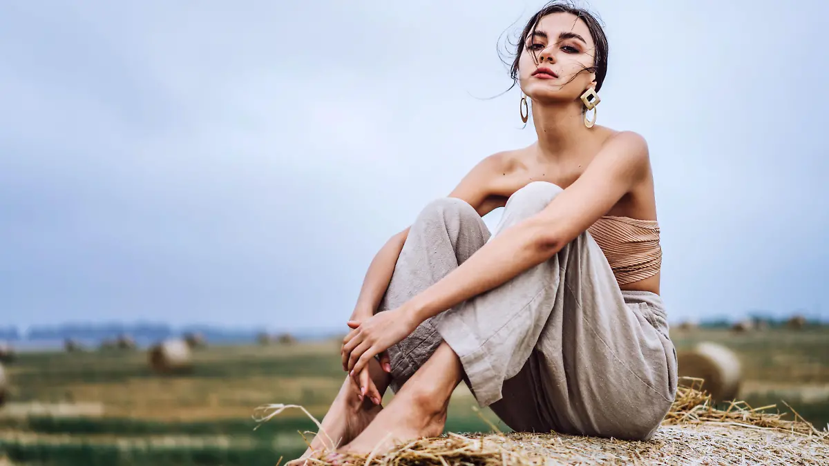 Brunette in linen pants and bare shoulders sitting on a hay bales in warm autumn day. Woman looking at camera. Behind her is a wheat field.
