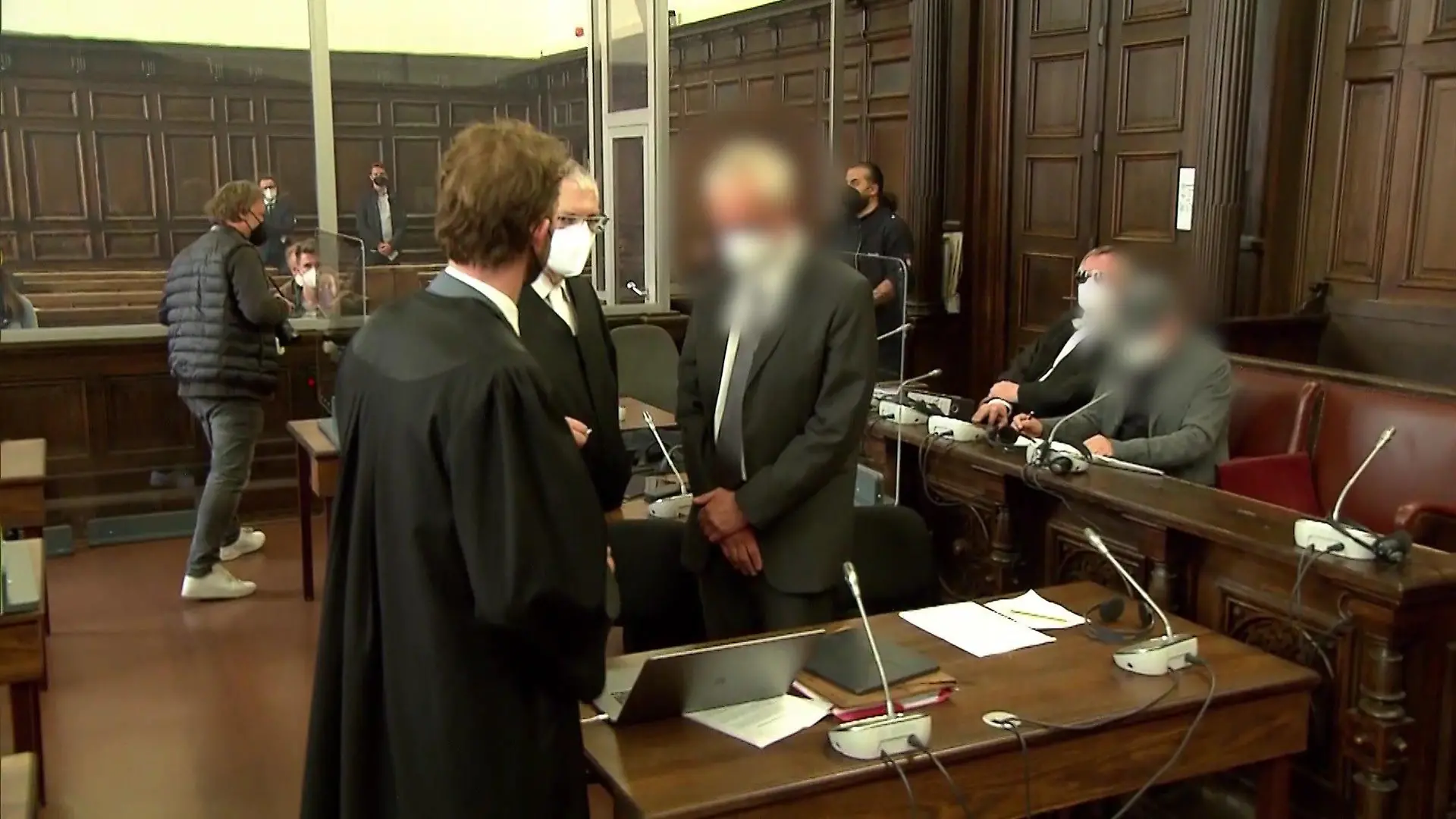 Die beiden Angeklagten kurz vor dem Prozess, umgeben von ihren Anwälten. Der eine sitzt bereits, während der andere noch steht.