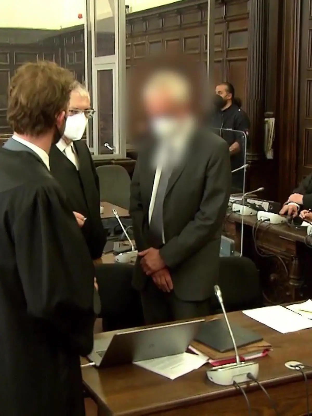 Die beiden Angeklagten kurz vor dem Prozess, umgeben von ihren Anwälten. Der eine sitzt bereits, während der andere noch steht.