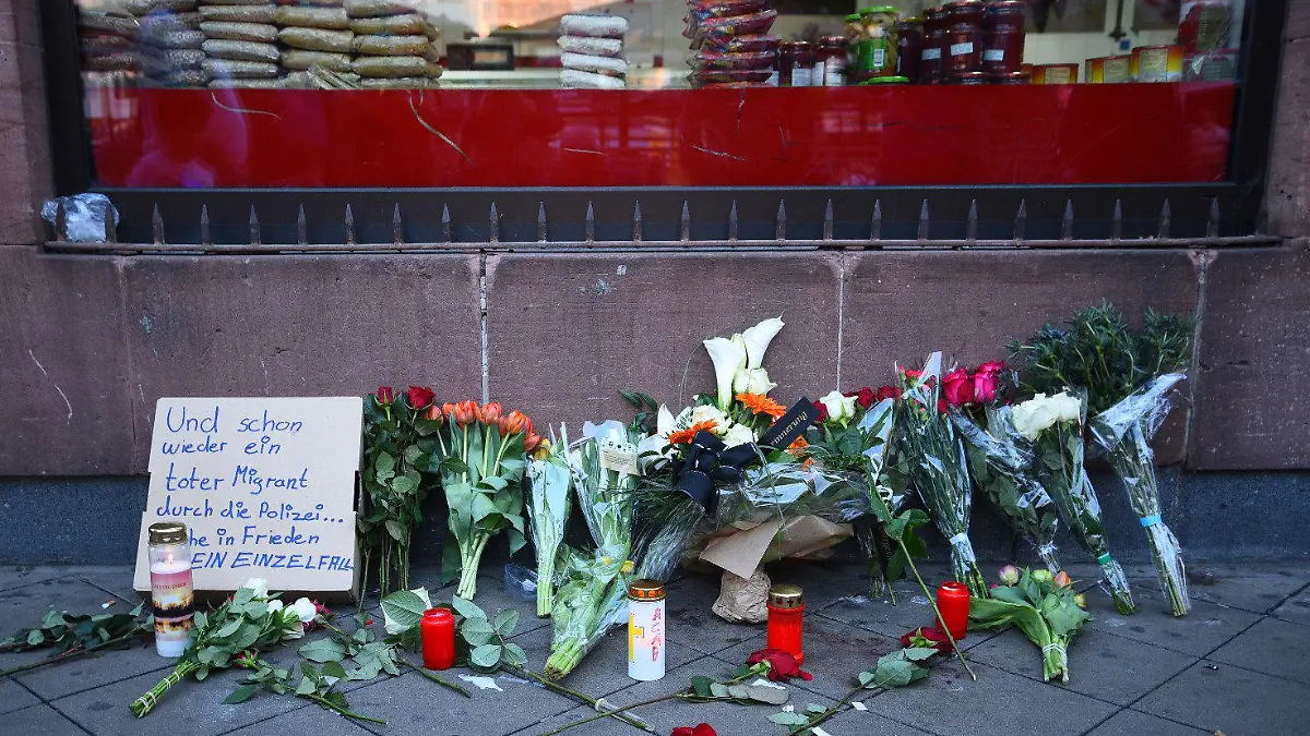 02.05.2022, Baden-Württemberg, Mannheim: Blumen liegen an dem Ort, an dem am Montag ein Mann nach einer Polizeikontrolle gestorben ist. Daneben steht ein Pappschild mit der Aufschrift «Und schon wieder ein toter Migrant durch die Polizei...». Die Leiche des Mannes wurde für weitere Untersuchungen beschlagnahmt, wie die Staatsanwaltschaft Mannheim und das Landeskriminalamt (LKA) Baden-Württemberg in Stuttgart mitteilten. Foto: René Priebe/PR-Video /dpa +++ dpa-Bildfunk +++