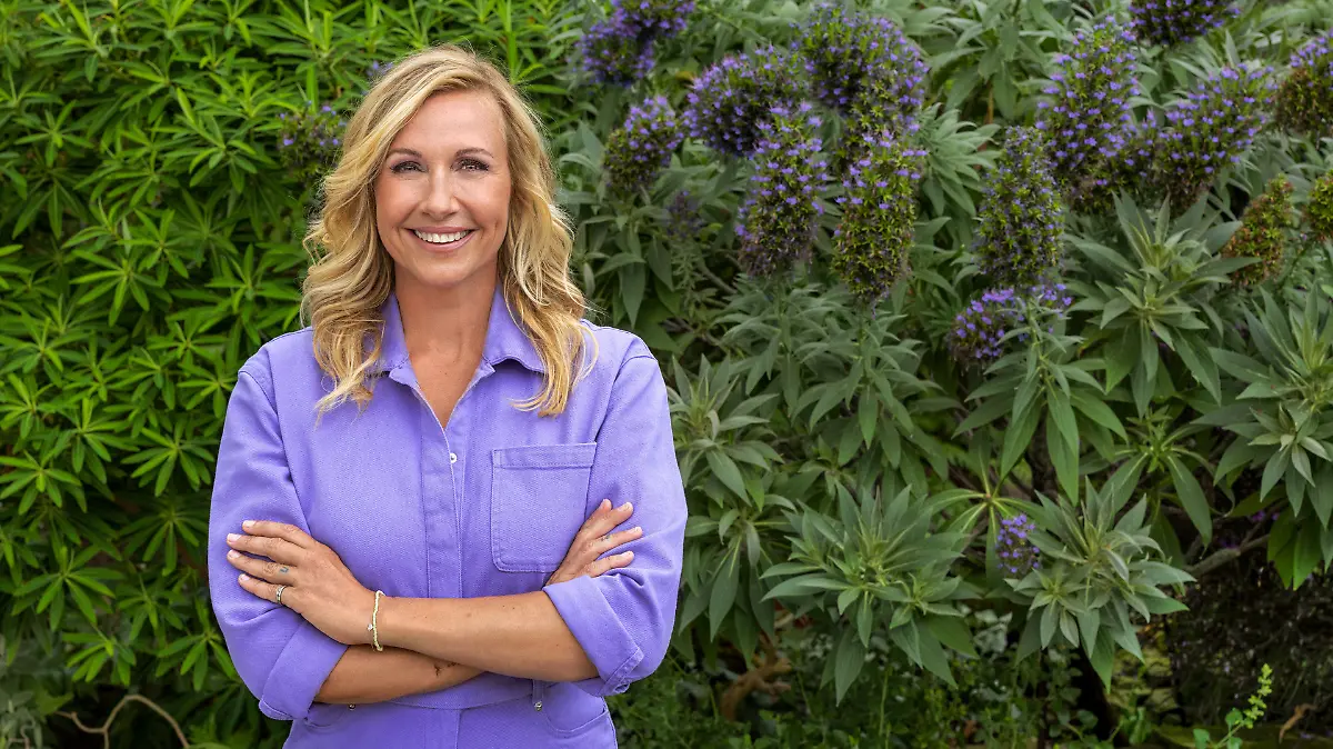 Andrea Kiewel steht in einem lila Hosenanzug vor einer Hecke.