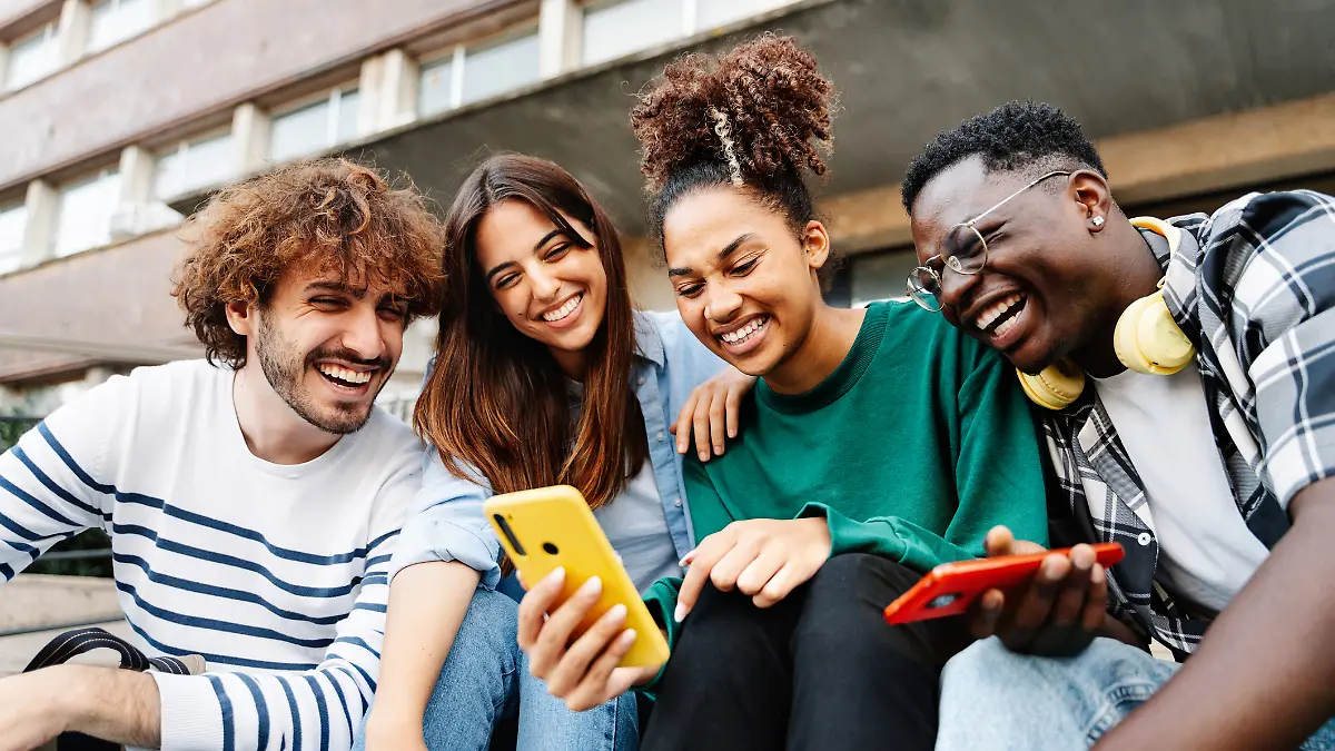 Zwei Männer und zwei Frauen sitzen lächelnd gemeinsam vor einer Universität und schauen auf ein Smartphone. 