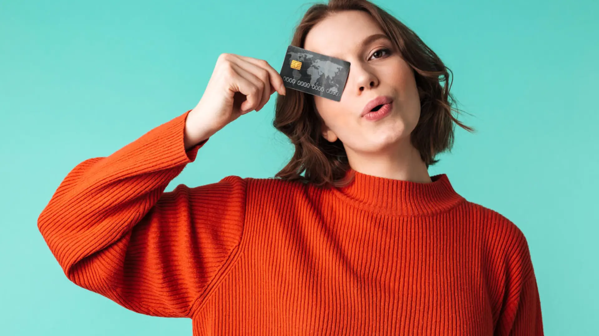Portrait of a pretty young woman dressed in sweater holding credit card at her face isolated over blue background