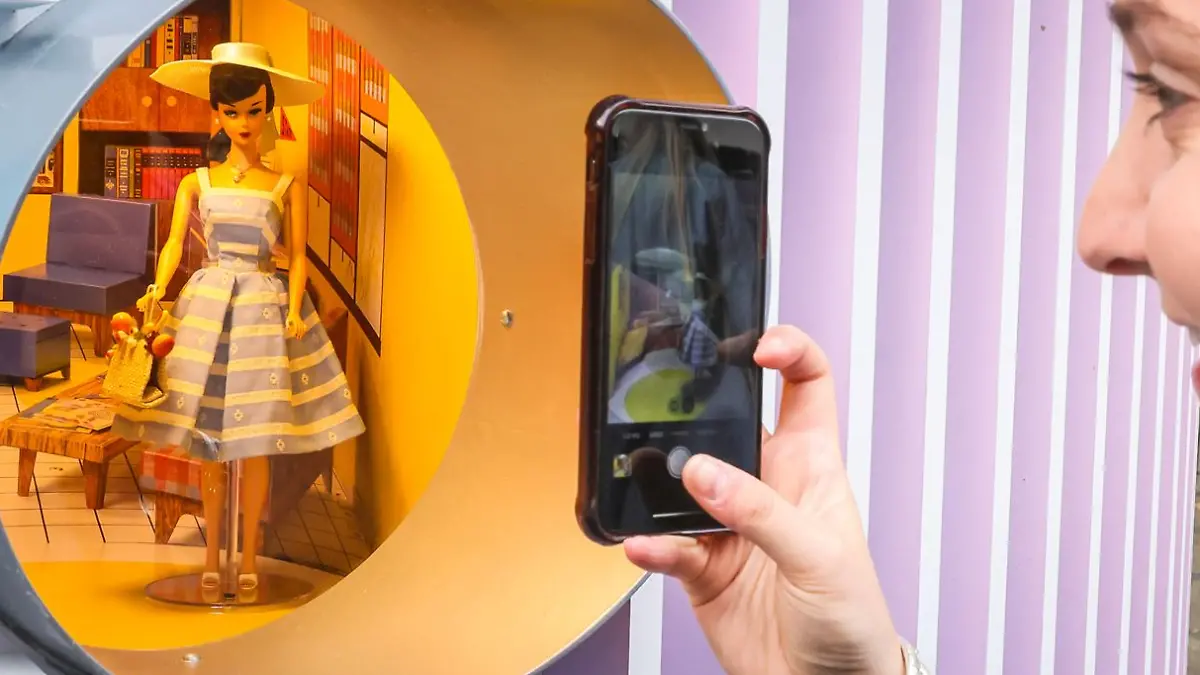 Eine 50er-Jahre Barbie steht hinter einem runden Fenster und wird von einer Frau im Profil mit Handy fotografiert.