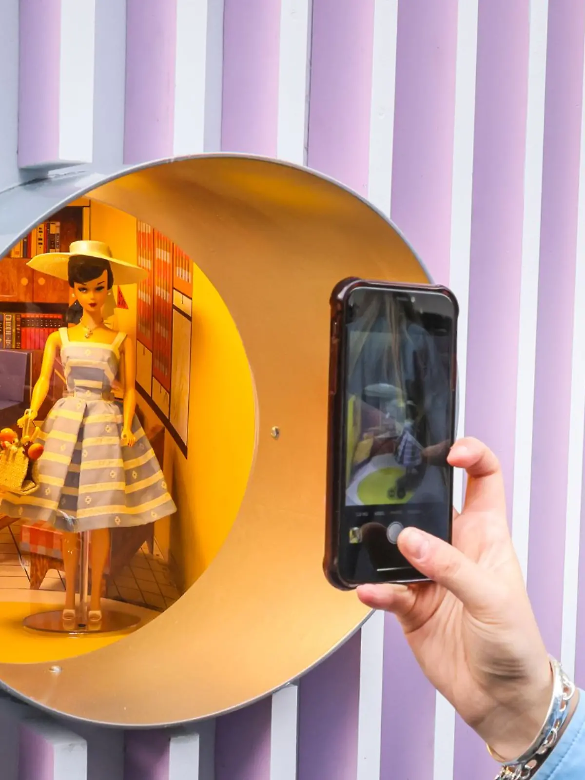Eine 50er-Jahre Barbie steht hinter einem runden Fenster und wird von einer Frau im Profil mit Handy fotografiert.