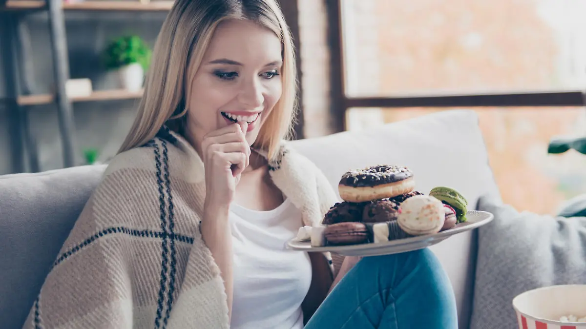 Portrait of beautiful emotional charming attractive sweet toothy woman sitting on sofa in living room, holding plate of donuts and macaroons, looking exciting satisfied